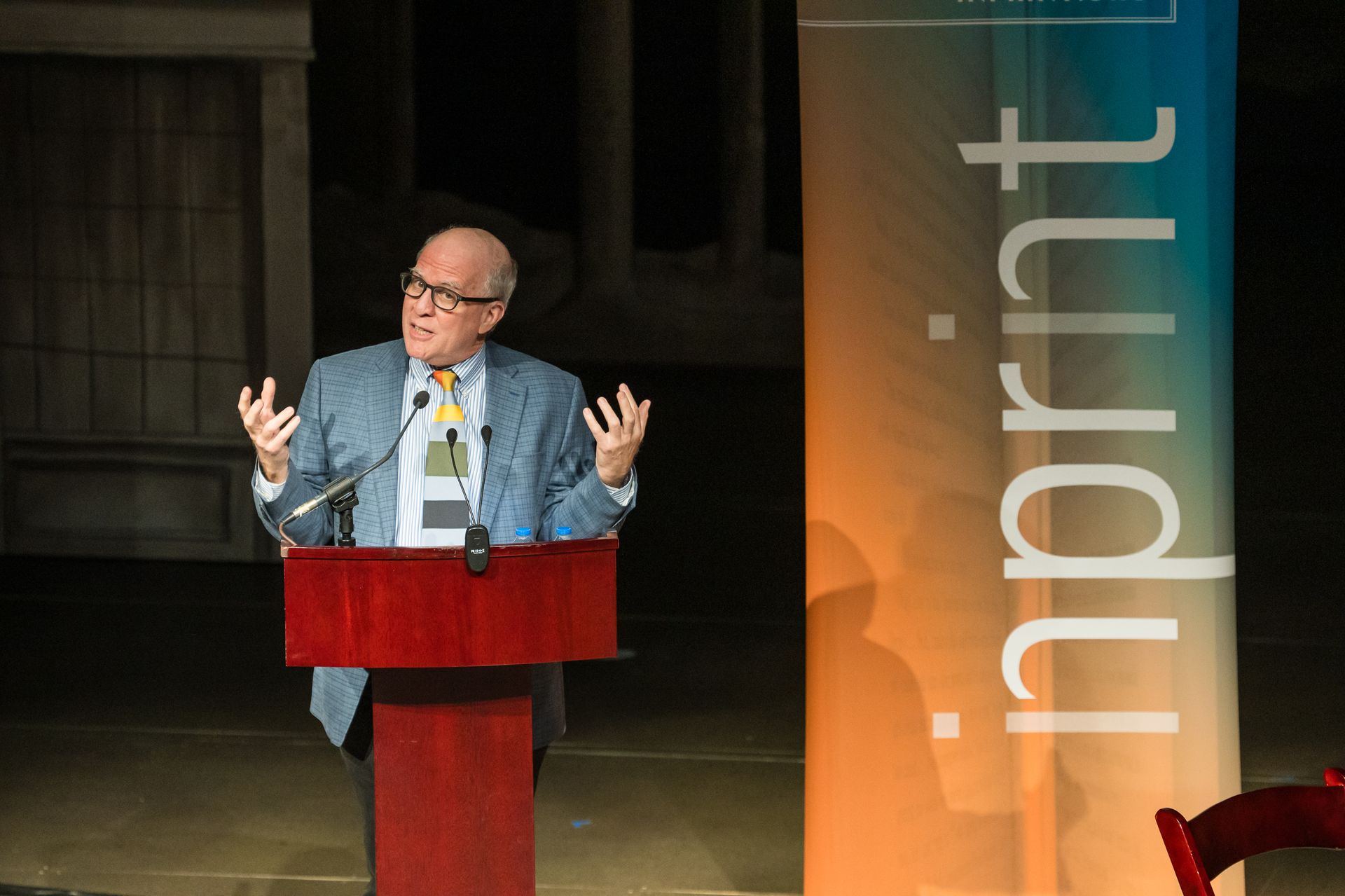 Man at a podium speaking, gesturing with hands. A banner with