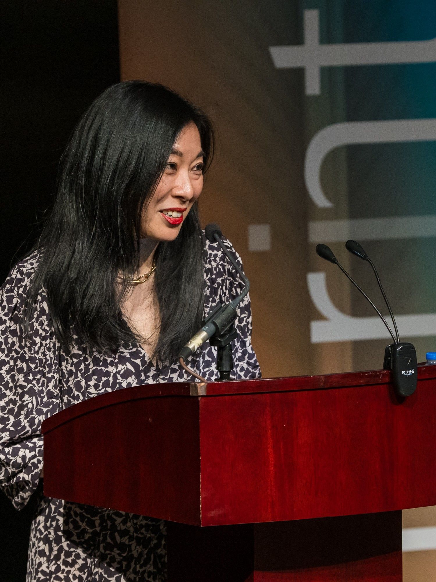 Woman speaking at a podium, wearing a patterned dress. Microphone and logo visible.