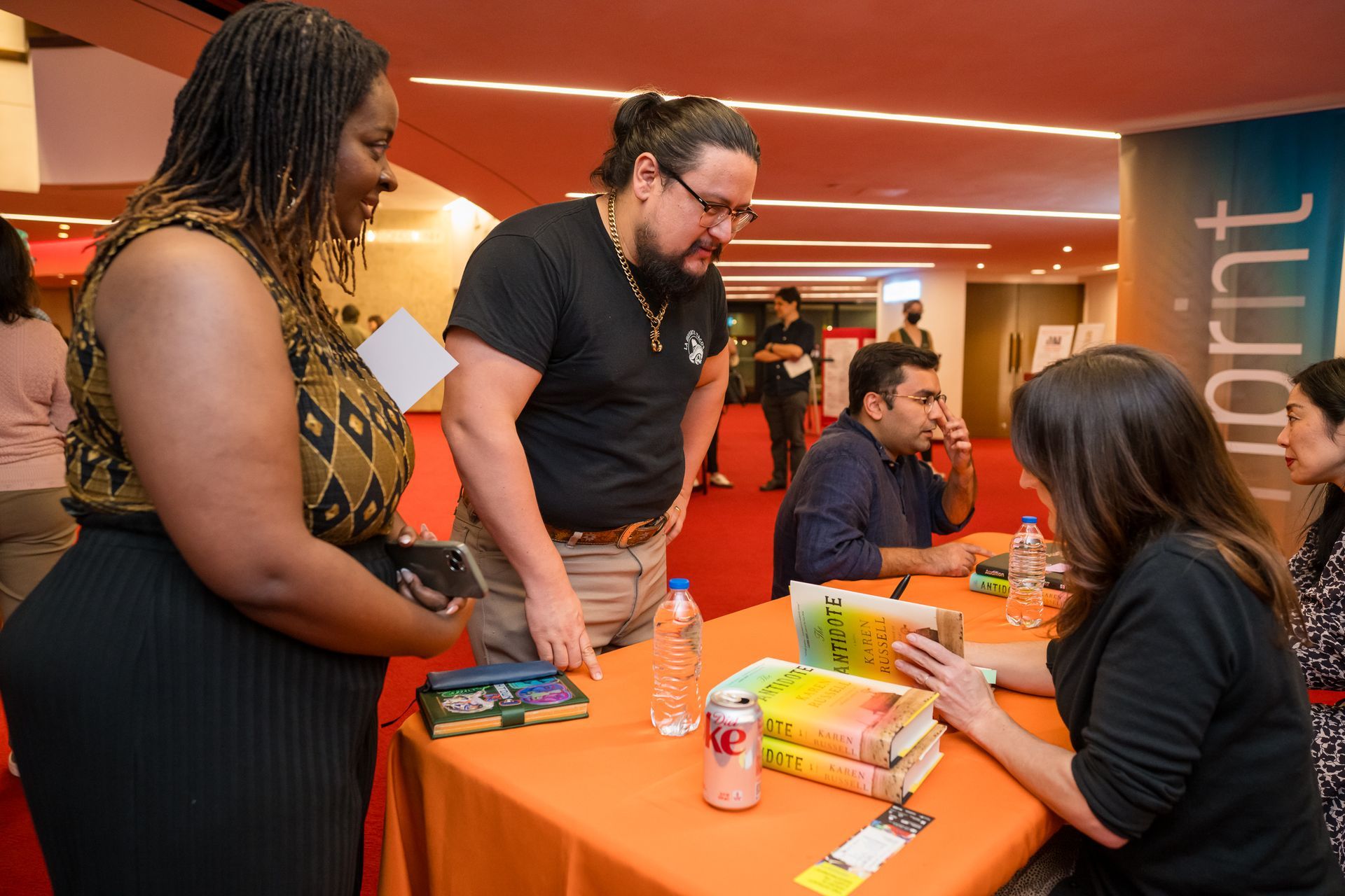 People at a book signing. Author seated, signing books on an orange table. Two people look on. Red carpet in a hall.