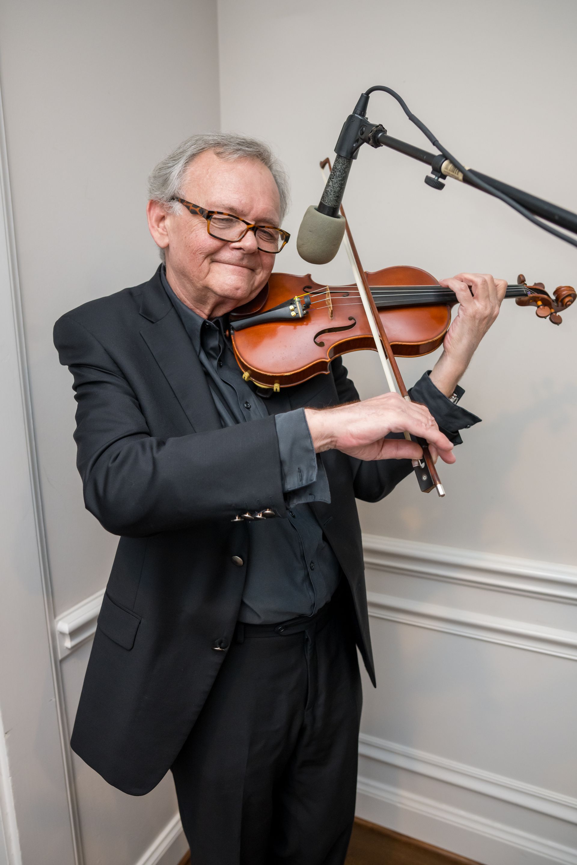 Man playing a violin into a microphone in a white-walled room, wearing glasses and a black suit.
