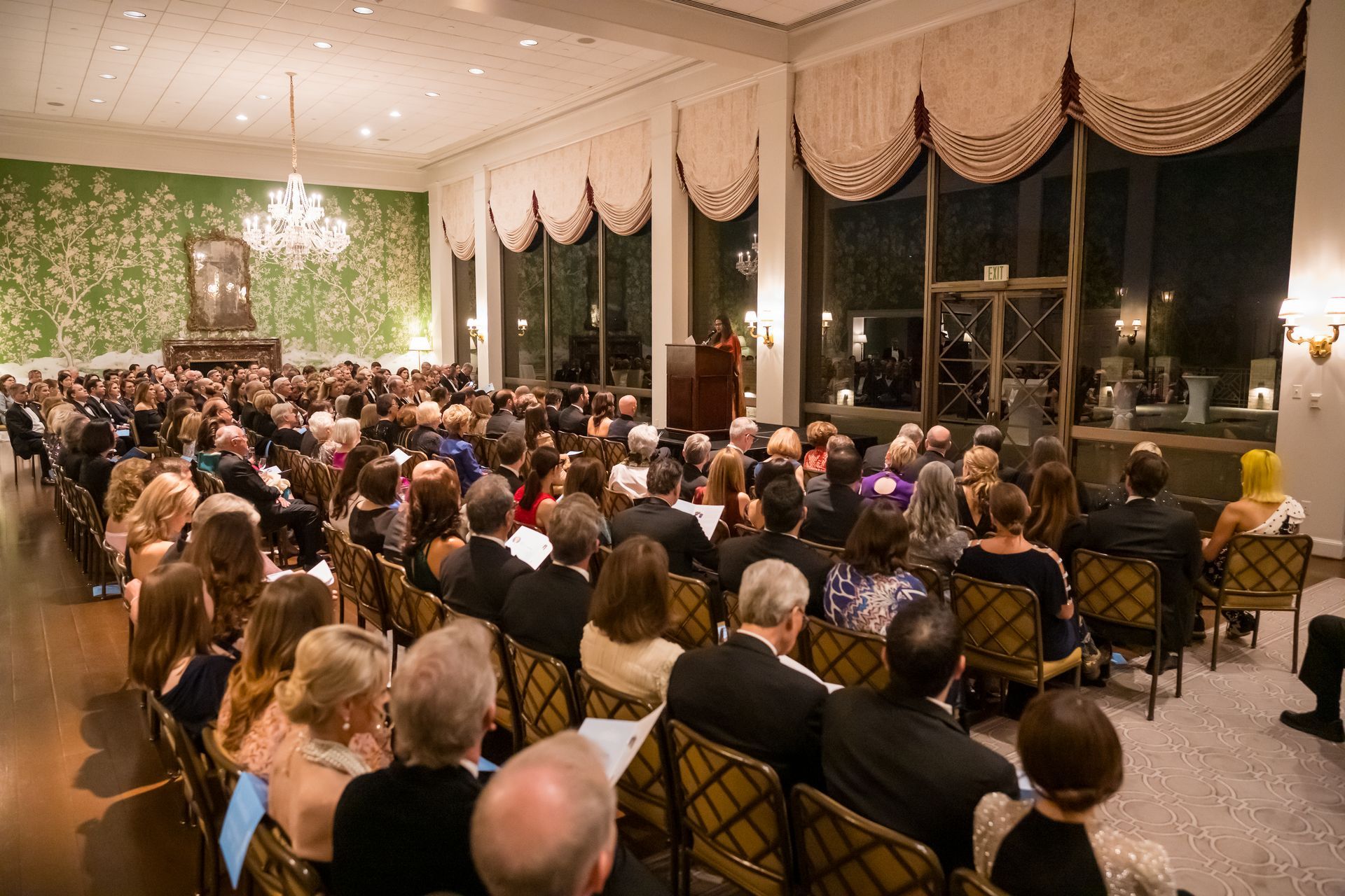Audience seated in a large room, listening to a speaker at a podium. Large windows. Chandelier.