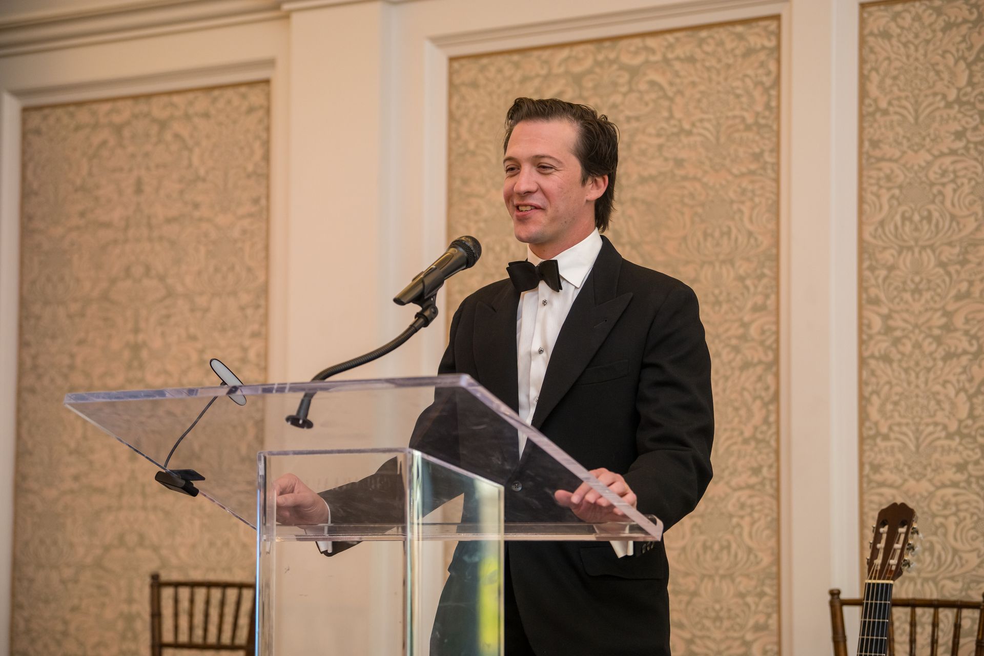 Man in a tuxedo speaking at a podium, smiling. Formal event setting with patterned wallpaper.