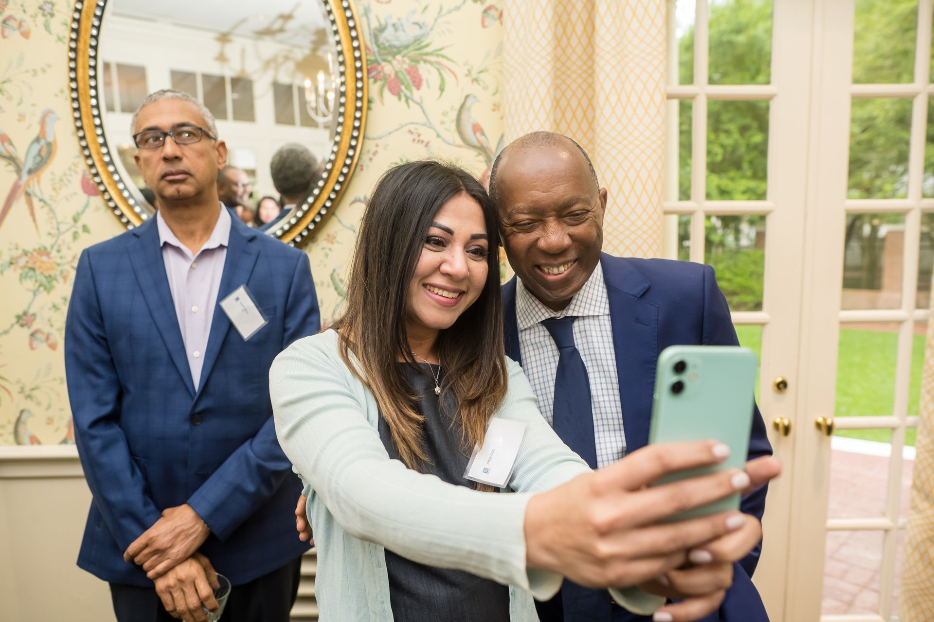 Woman taking selfie with a man, another man looks on. They are in a room with floral wallpaper and a mirror.
