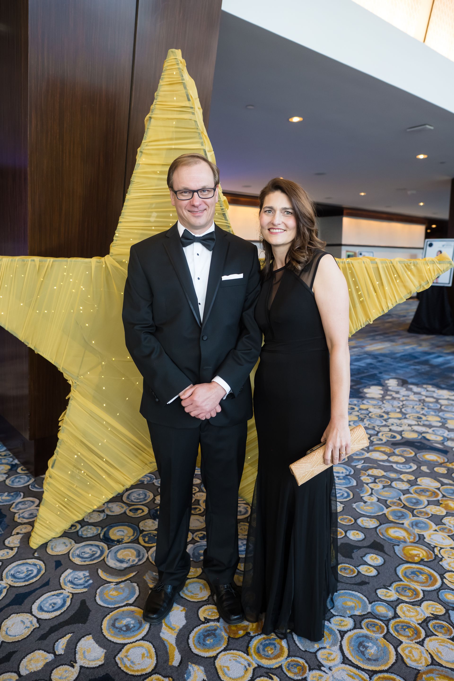 A couple poses in formal attire next to a large gold star decoration.