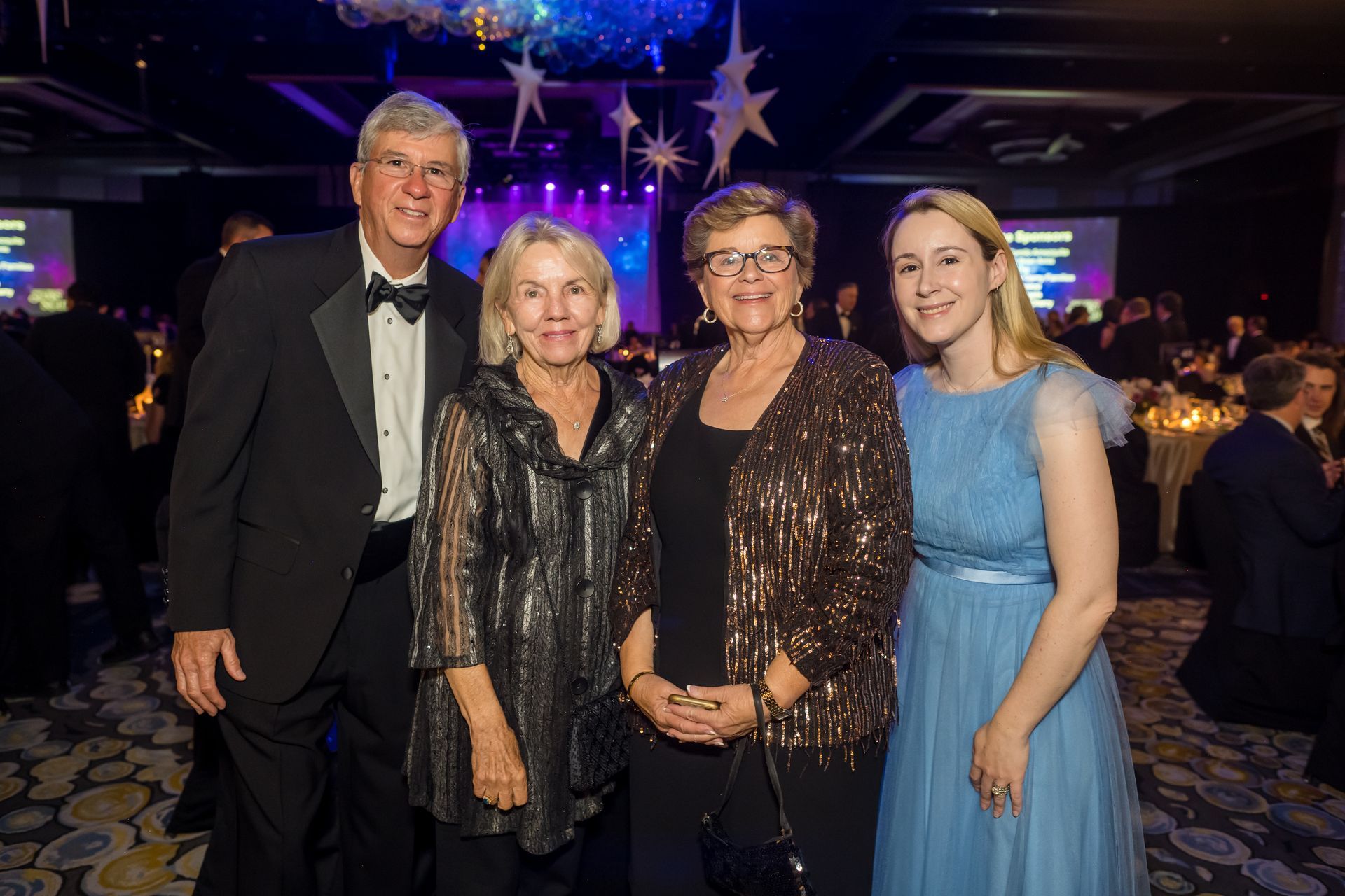 Four people smiling at a gala. Man in tux, three women in festive attire, indoors, tables set in background.