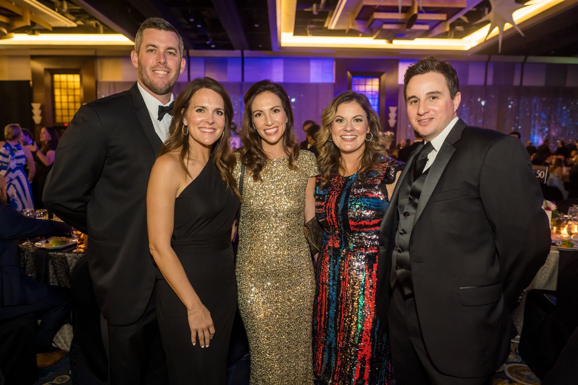 Five people in formal attire at an event, posing and smiling. Background features a lit room with tables.