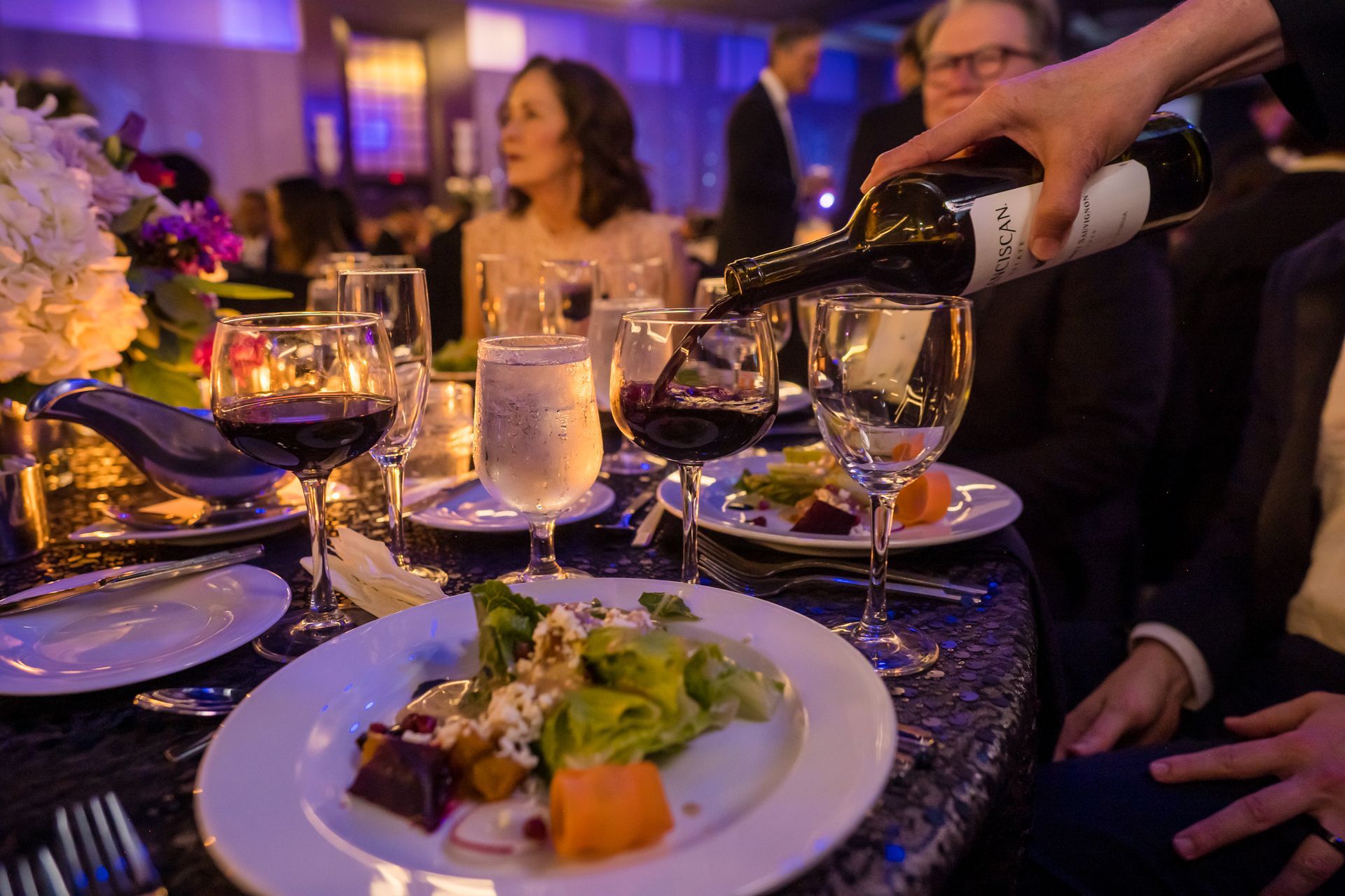 A hand pouring red wine into a glass at a formal event, with food and people at the table.