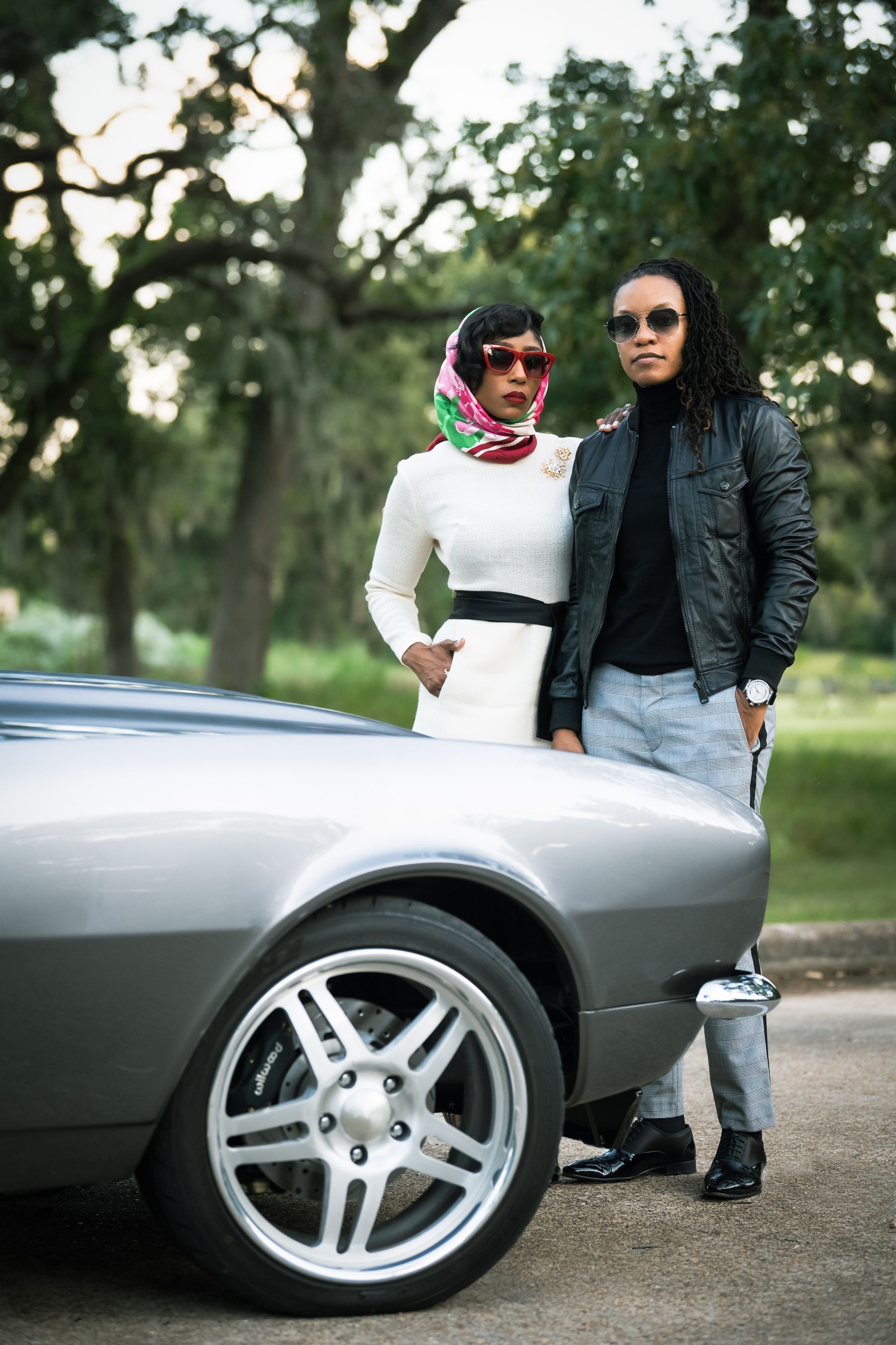 Two people in stylish attire pose by a silver classic car.