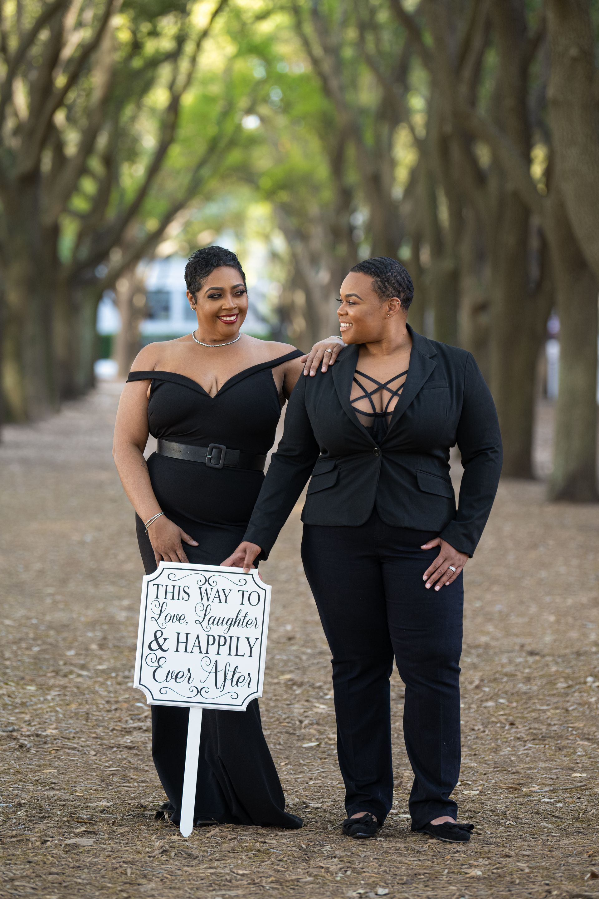 Two people in black formal attire hold a sign, smiling at each other, in a tree-lined walkway.