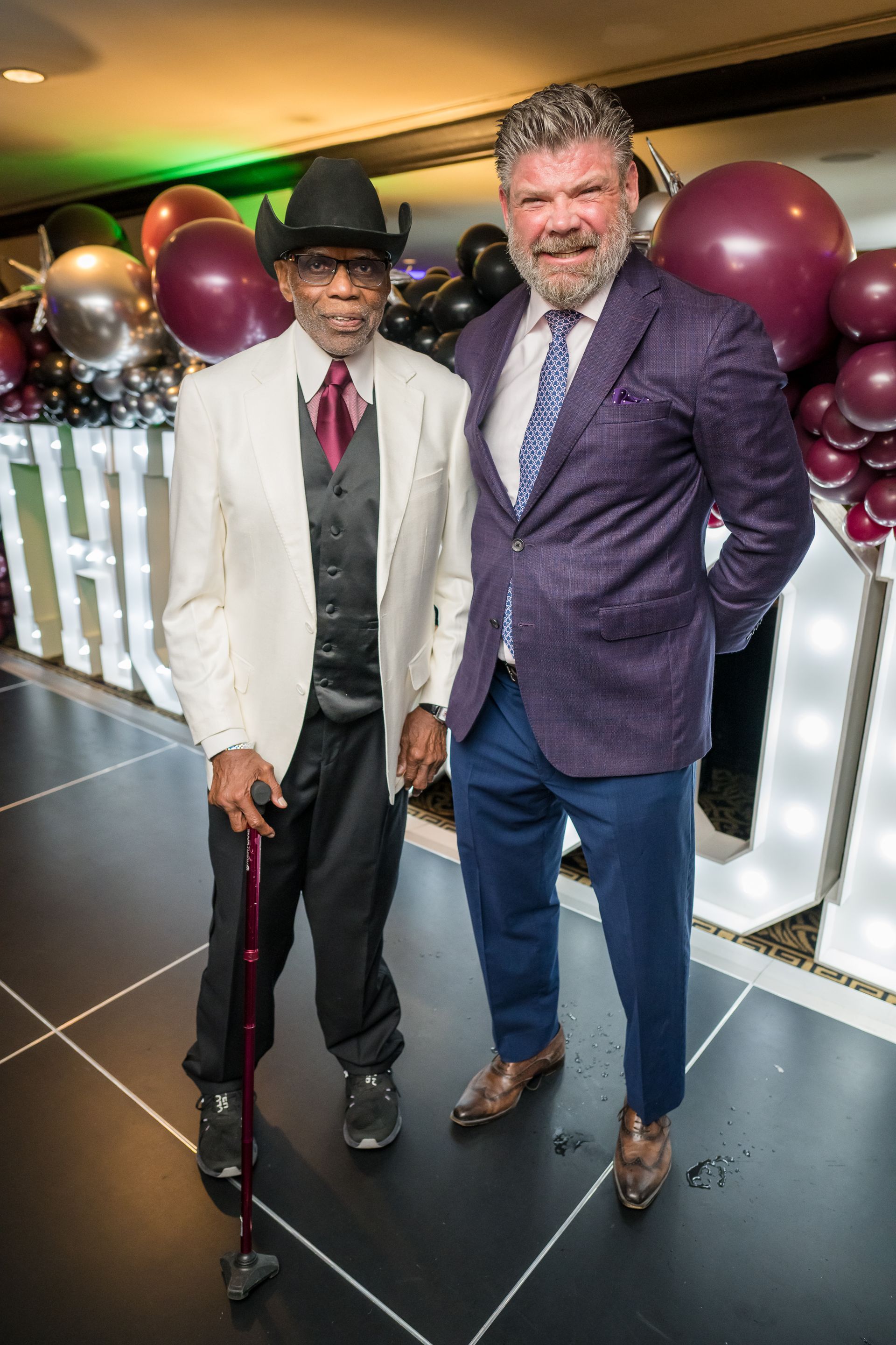 Two men pose together indoors. One in a white suit and hat, using a cane. The other in a purple blazer and blue pants.