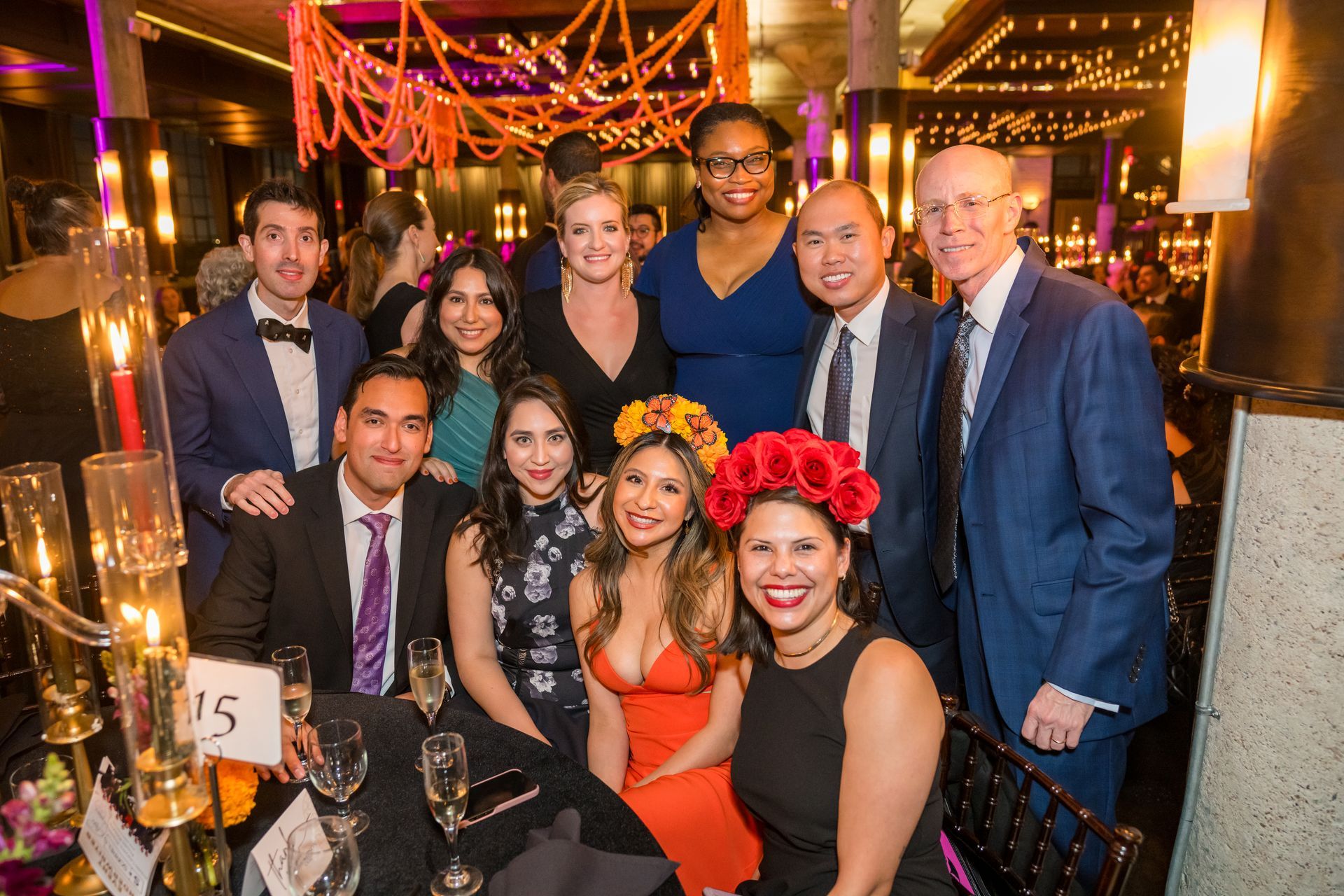 Group of people posing at a formal event, some wearing floral headbands. Table with candle holders.