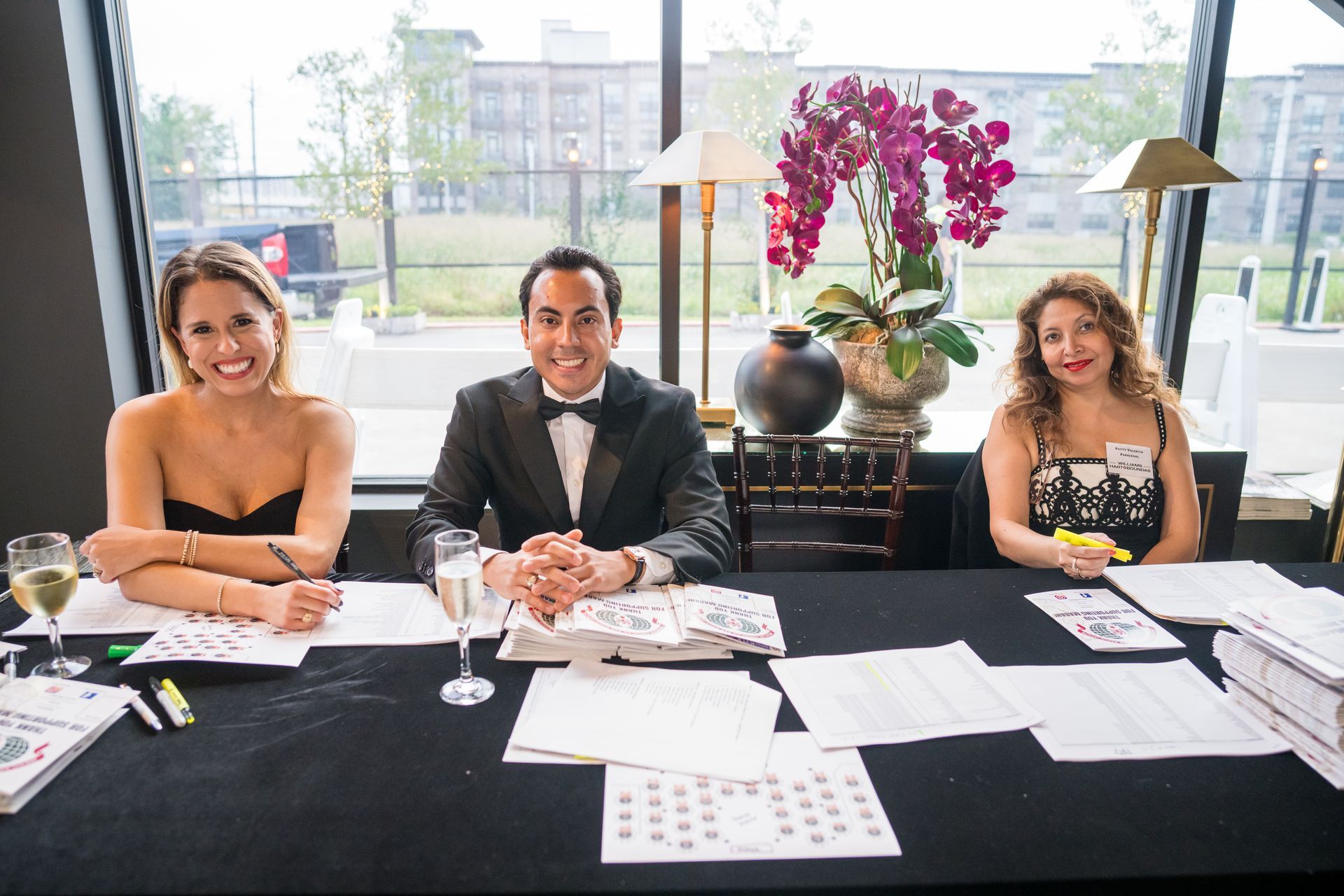 Three people at a registration table. Smiling, they appear to be at an event. Black tablecloth, window in background.