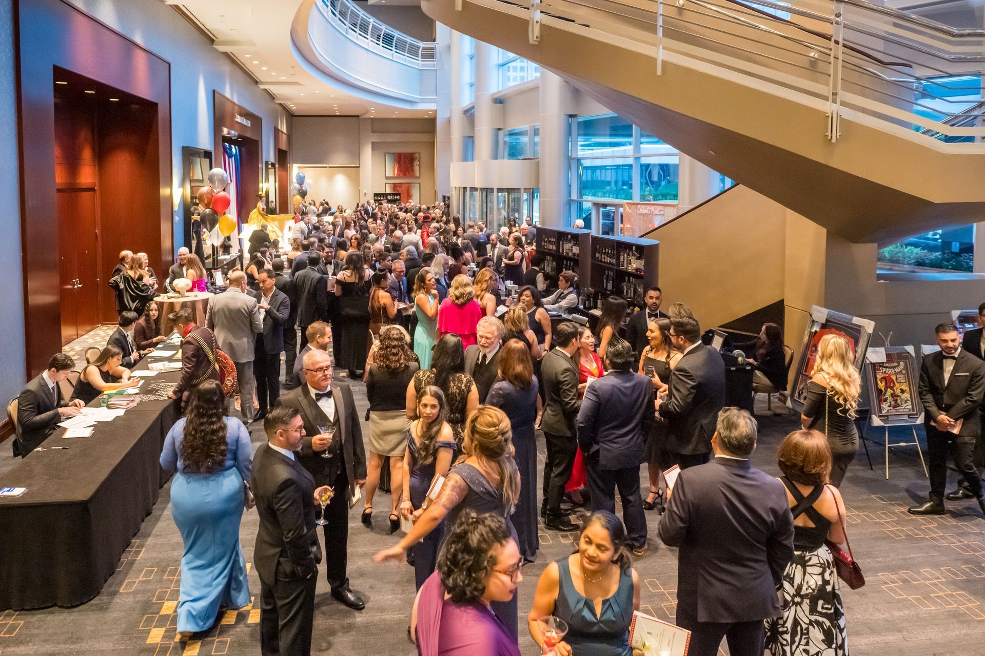Large group of formally dressed people at an event in a spacious lobby with stairs and a balcony.