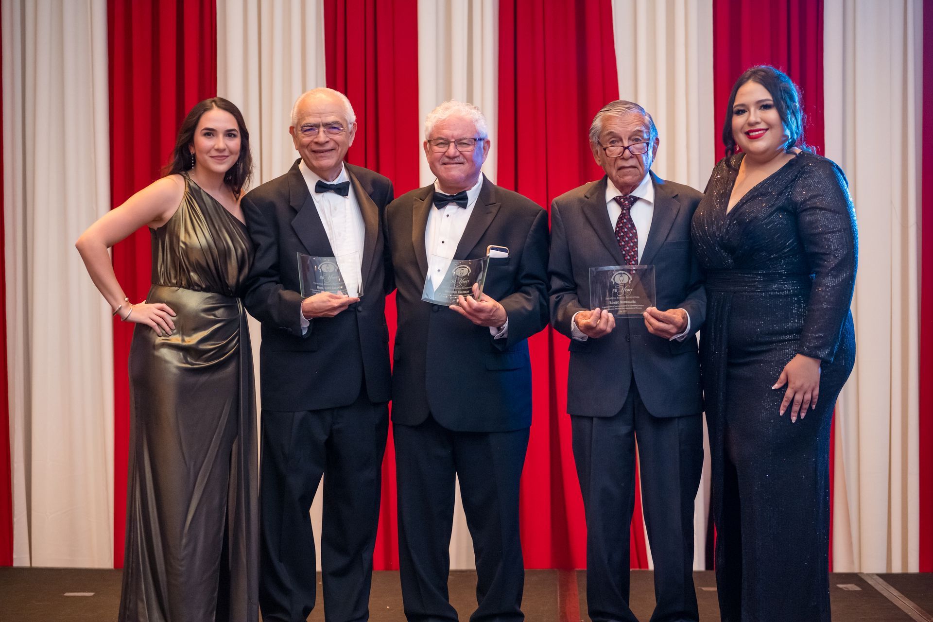 Five people, two women and three men, holding awards, posing in front of a red and white curtain.
