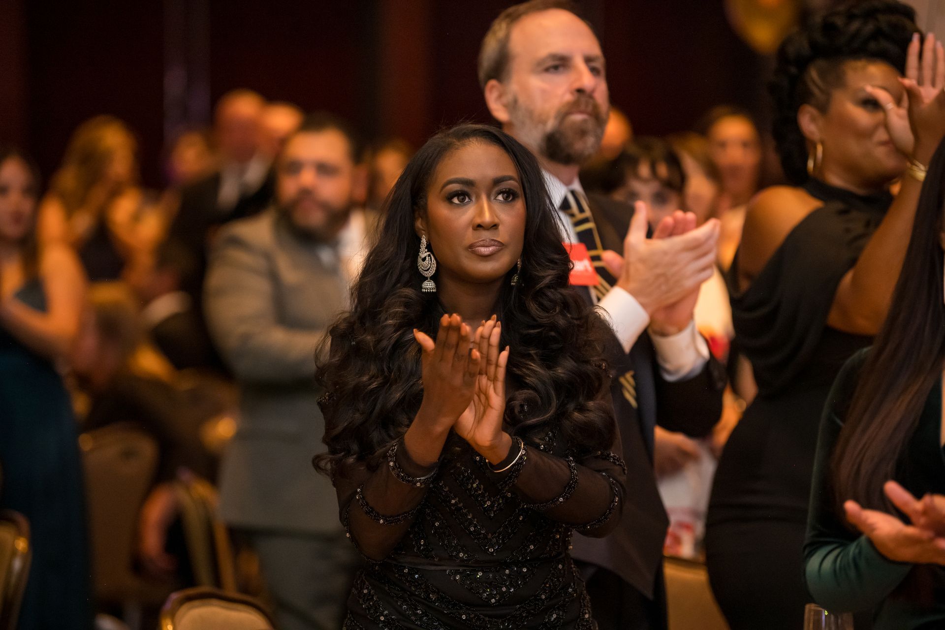 Woman in black dress applauding, at a gala event. People are visible, clapping, in a hall.