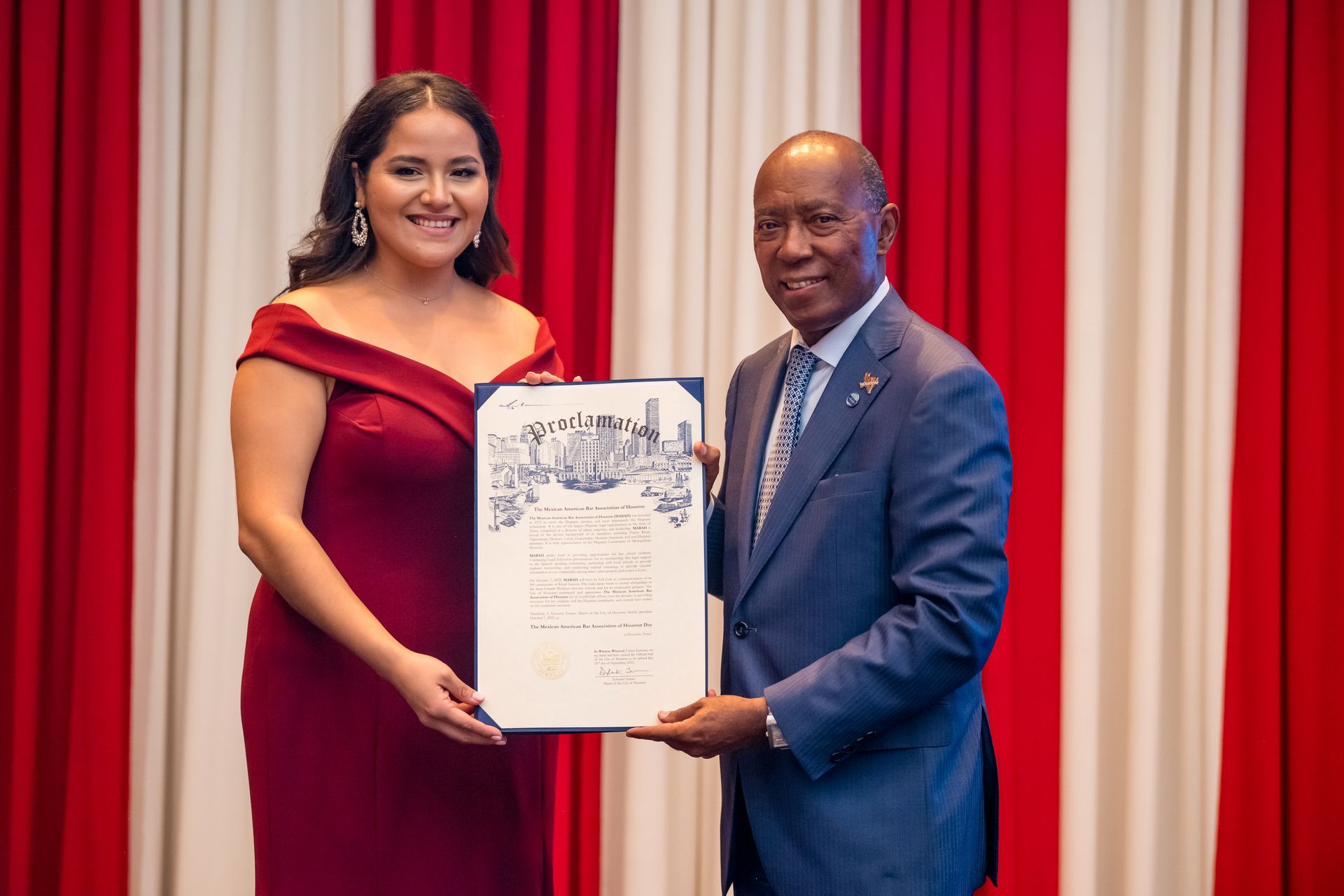 Woman in burgundy dress and man in a suit hold a framed document in front of red and white curtains.