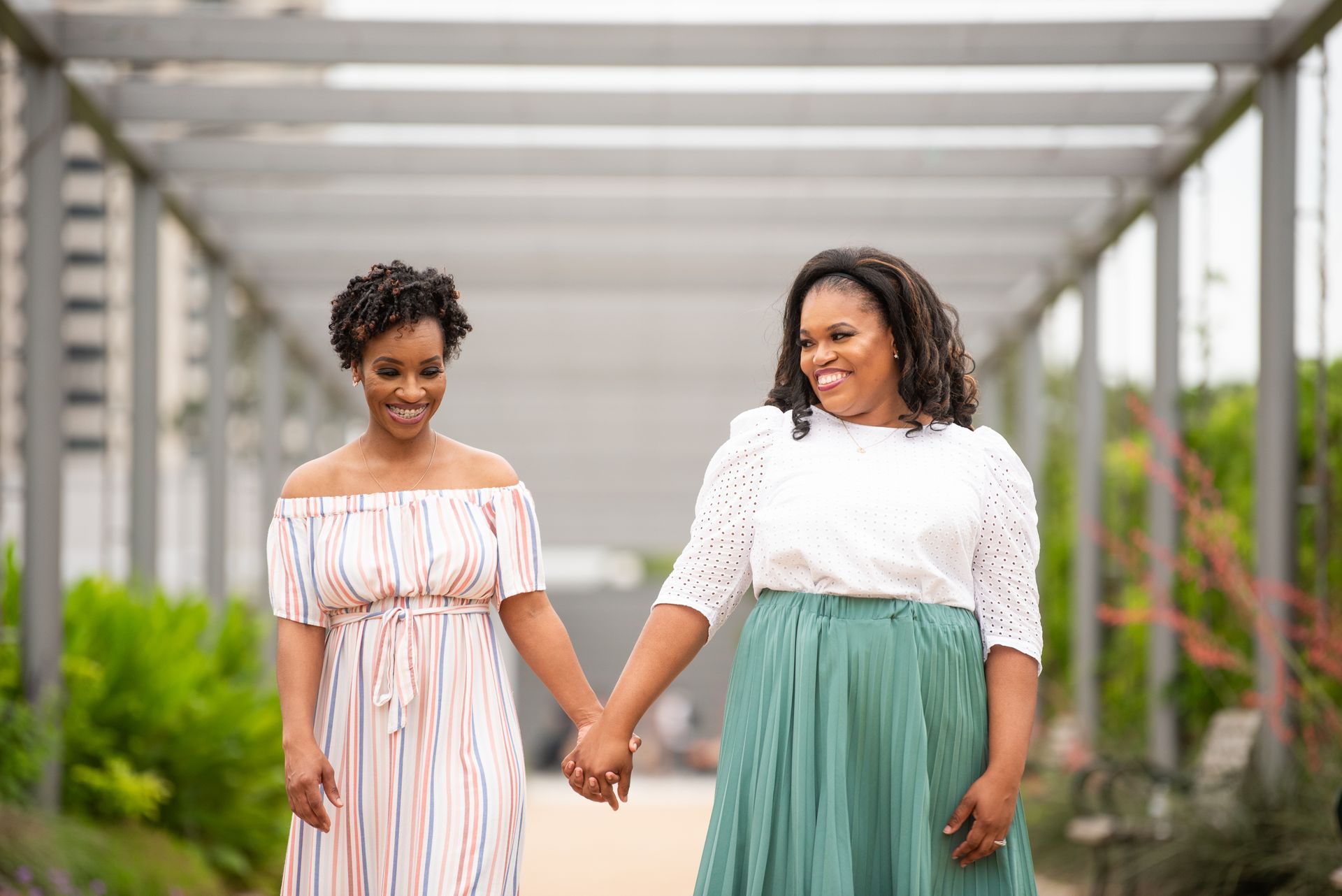 Two women holding hands, walking along a pergola-covered path.