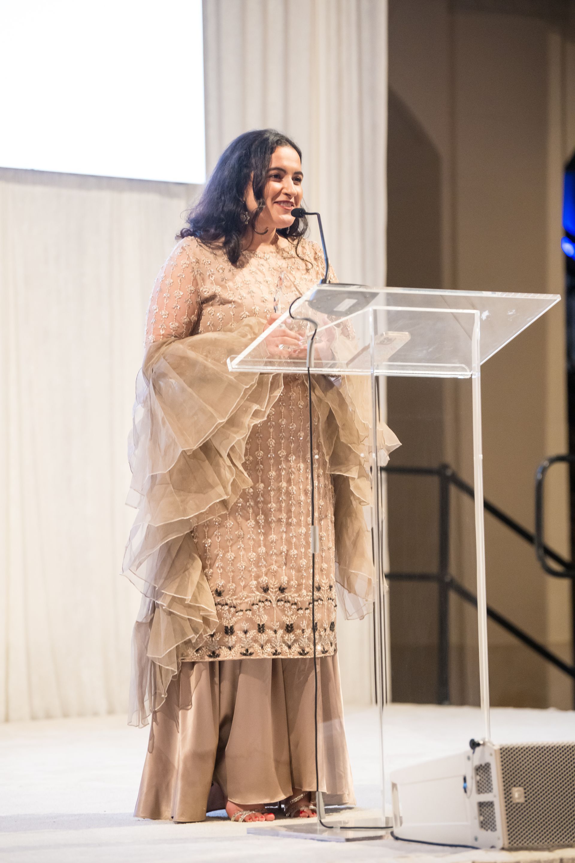 Woman in a tan dress speaks at a podium; stage setting.