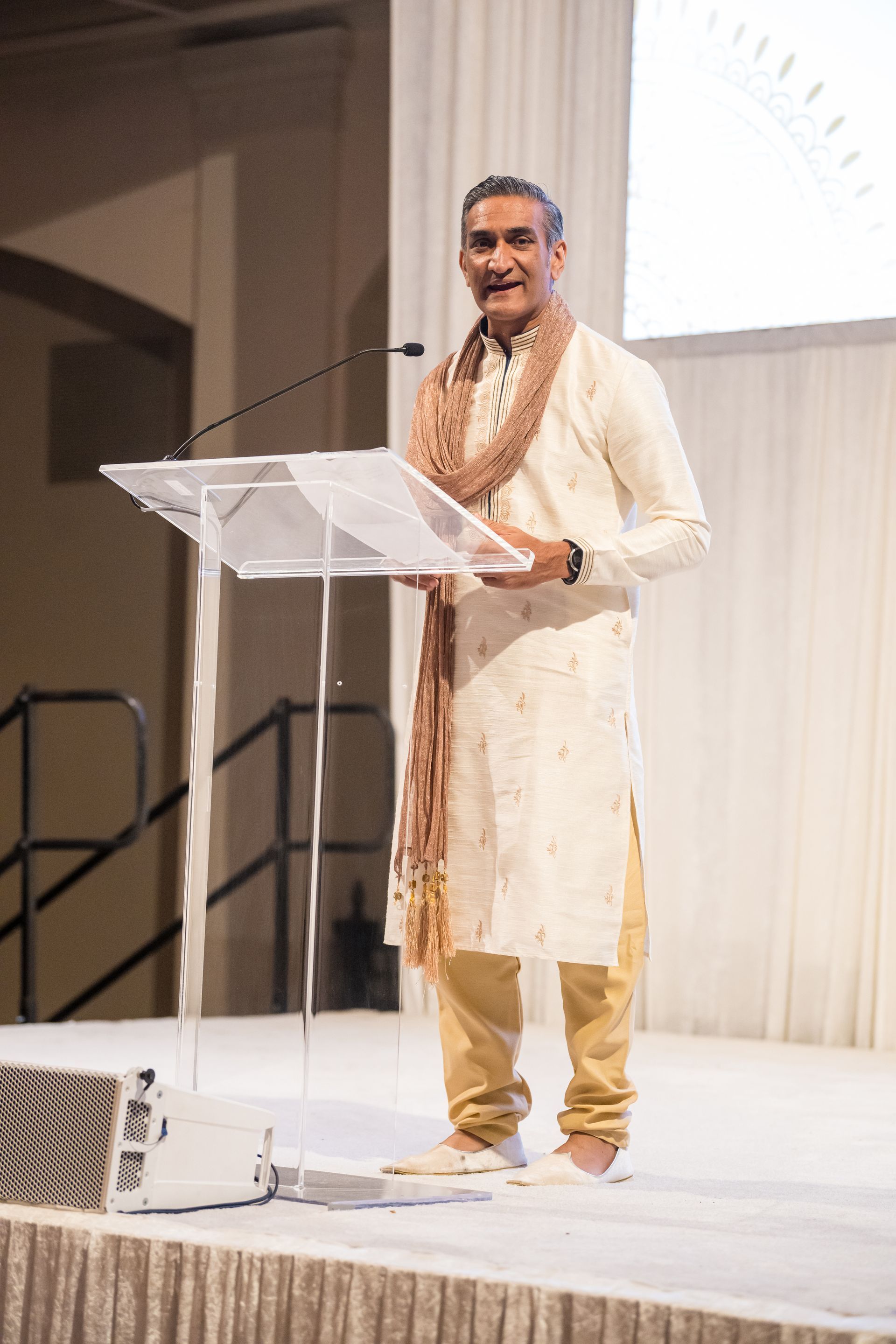 Man in traditional Indian attire giving a speech from a podium. Light background.