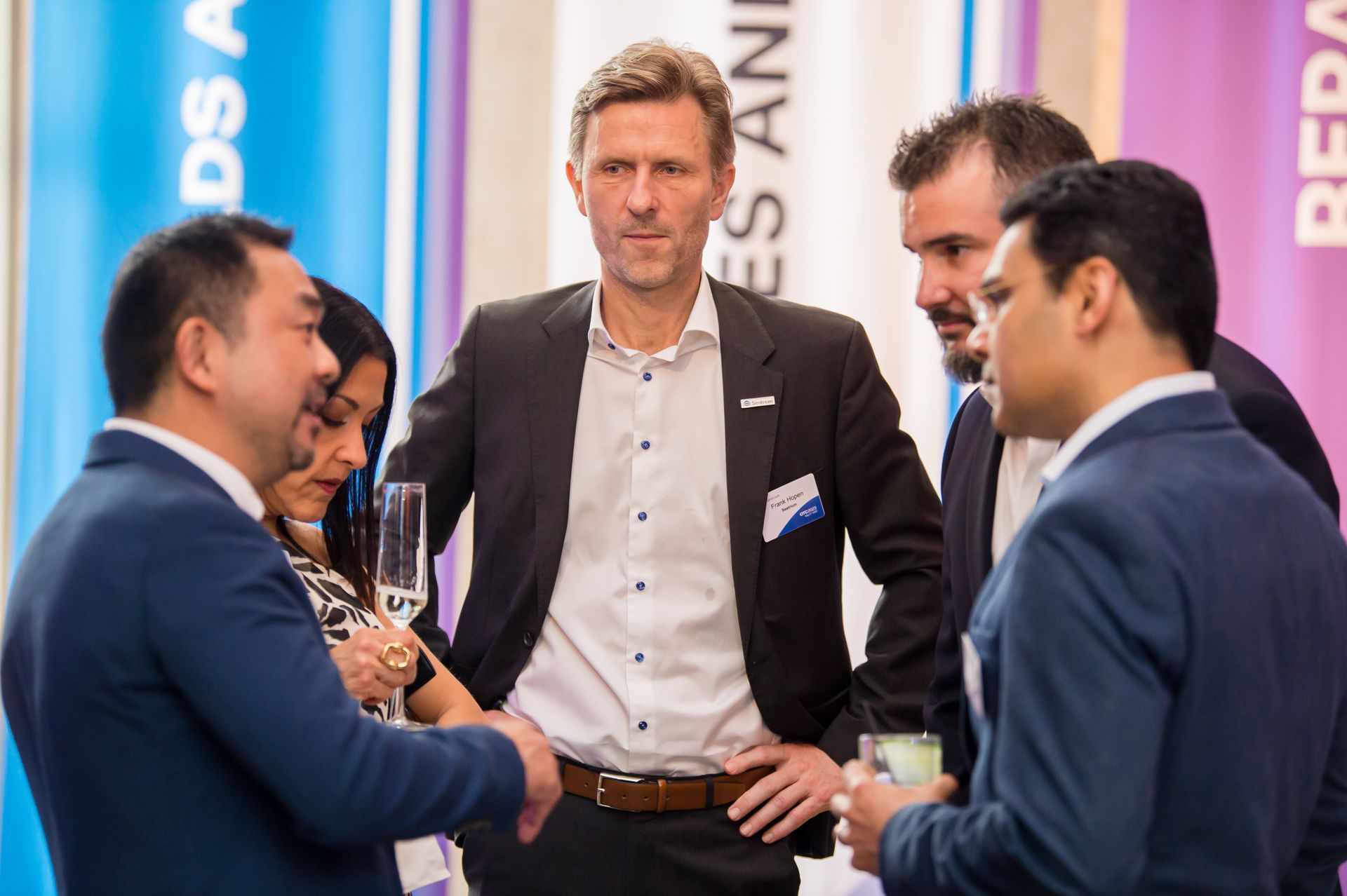 Group of people in suits talking at an event; indoor setting with banners.