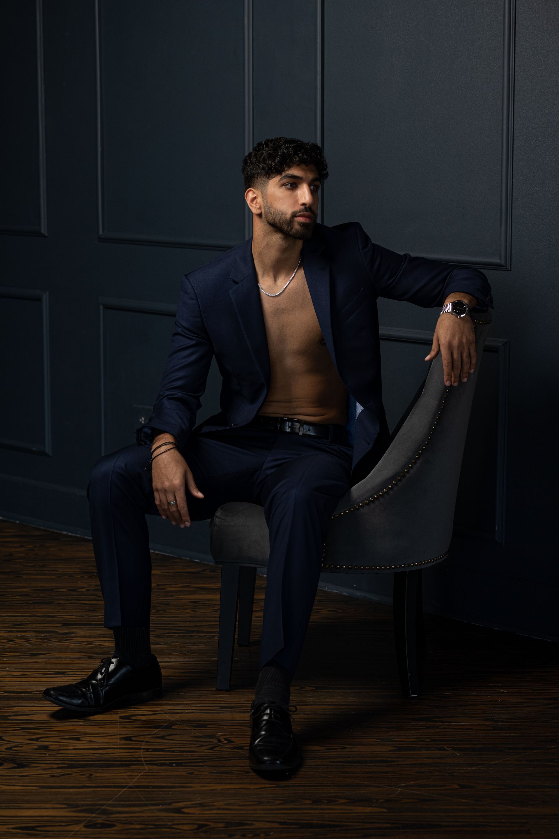 Man in blue suit, unbuttoned, seated, looking away. Dark background.