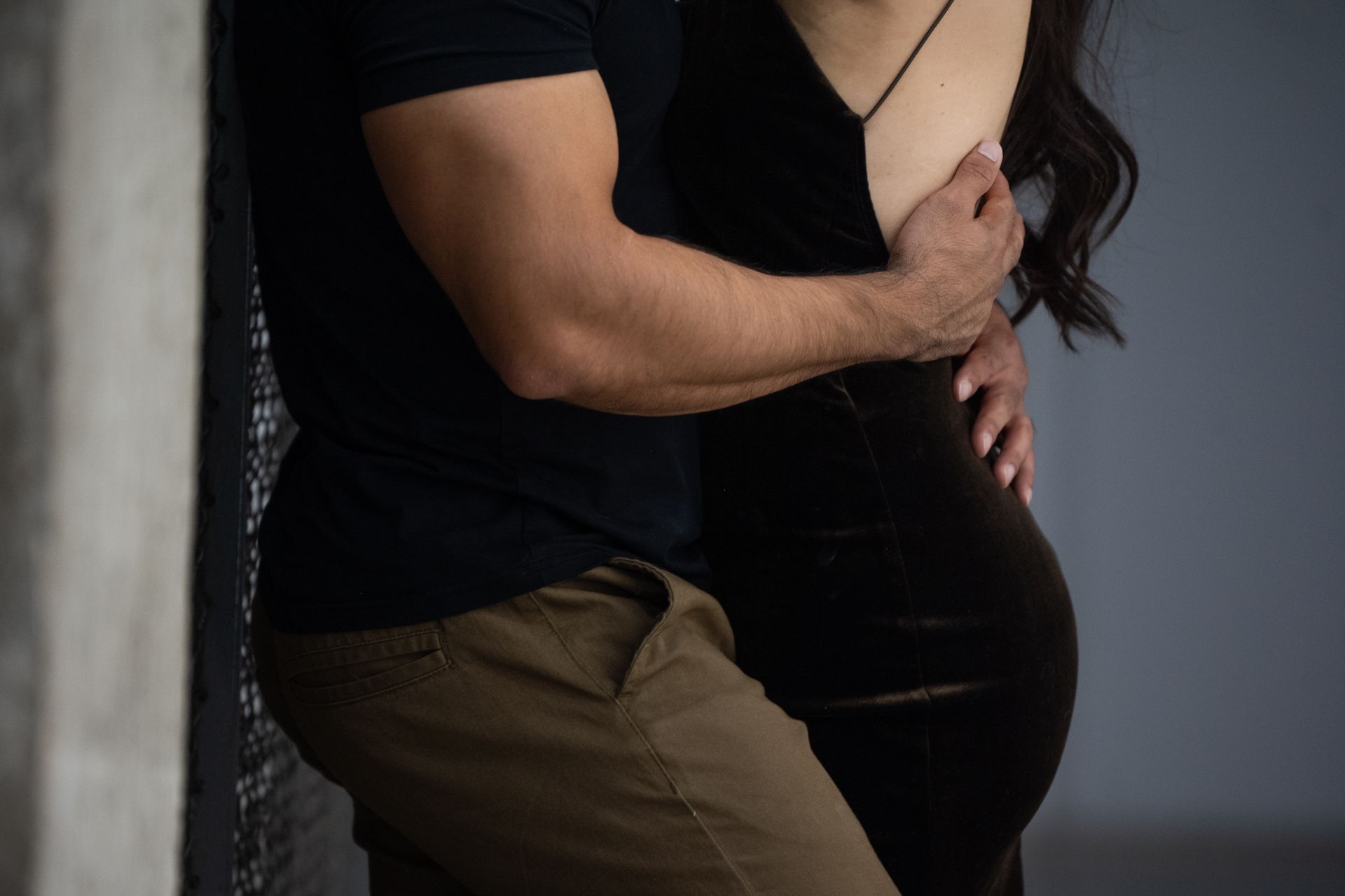 Man in black shirt and khaki pants embraces person in black dress against a wall.