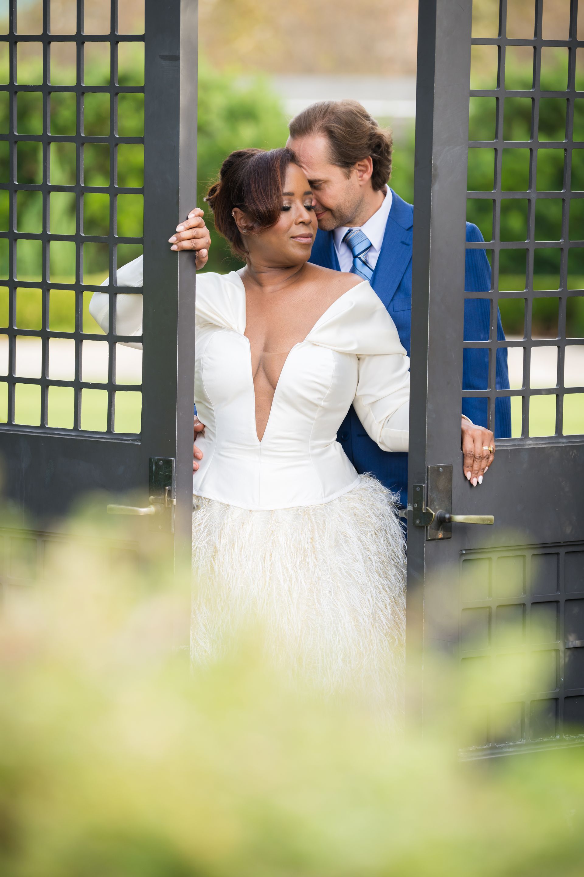 Couple, bride in white gown, groom in blue suit, posing at a black gate in a garden.