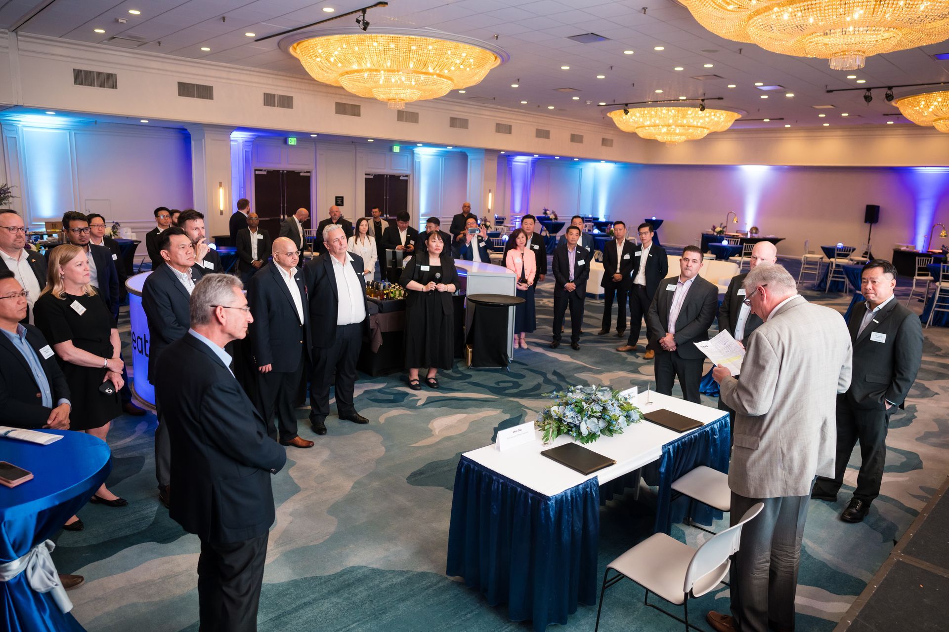 People gathered in a ballroom for an event. Several stand near a table with paperwork. Blue and gold accents throughout.