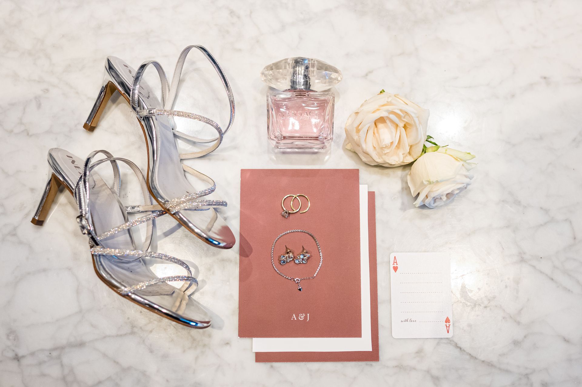 Silver high heels, perfume, roses, and wedding invitations on a marble surface.