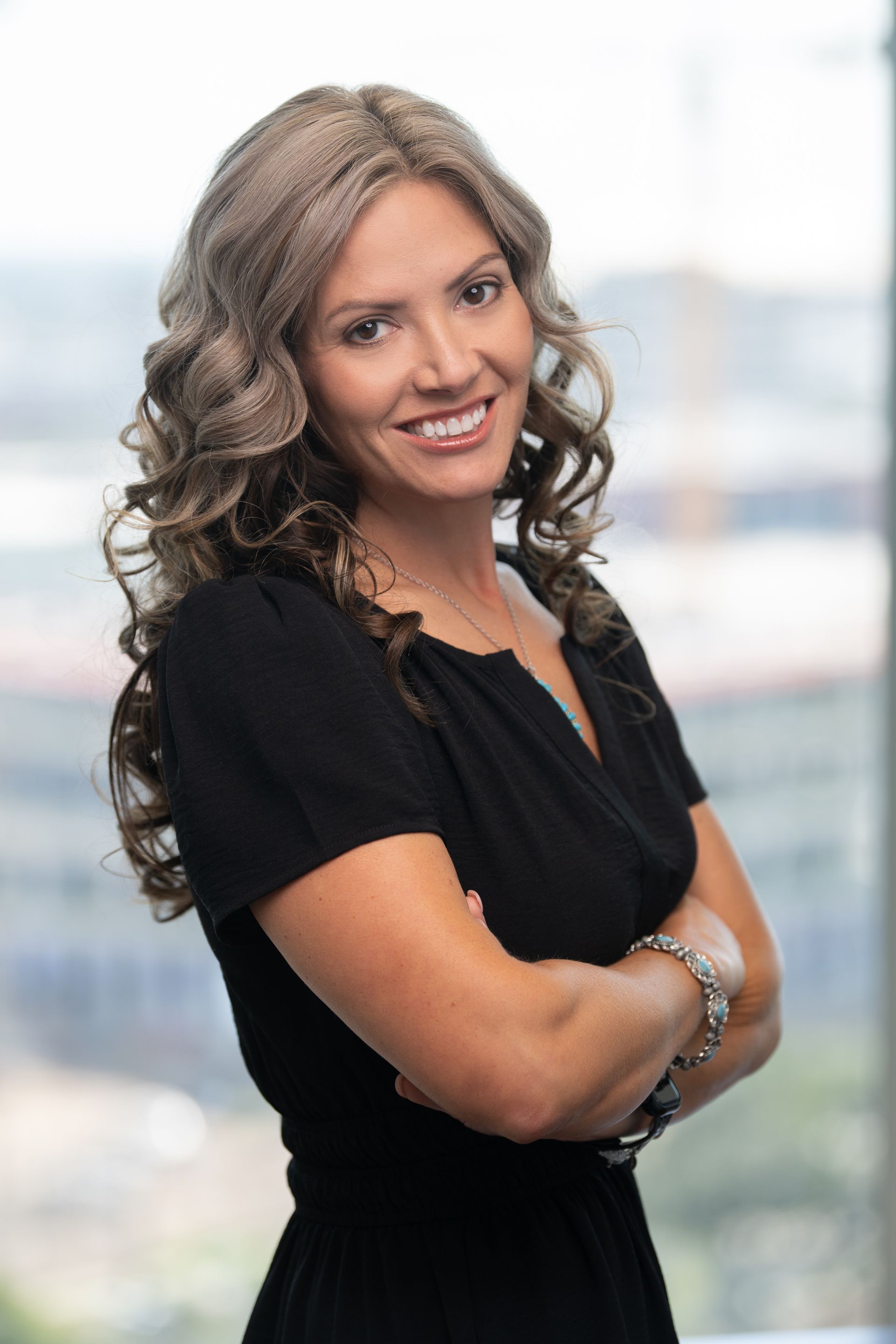 Woman with curly gray hair, black top, arms crossed, smiling in front of a window.