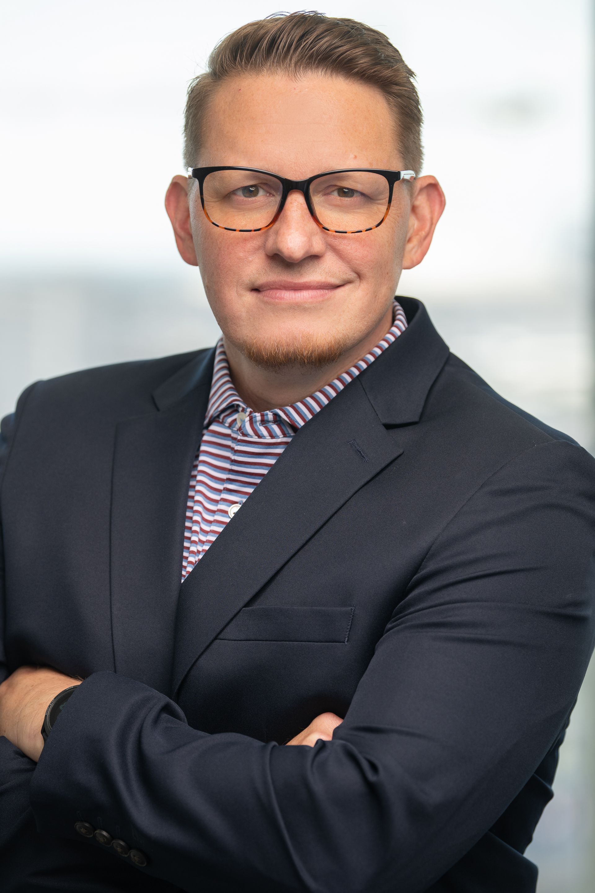 Man in glasses and blazer with arms crossed, smiling, in front of a window.