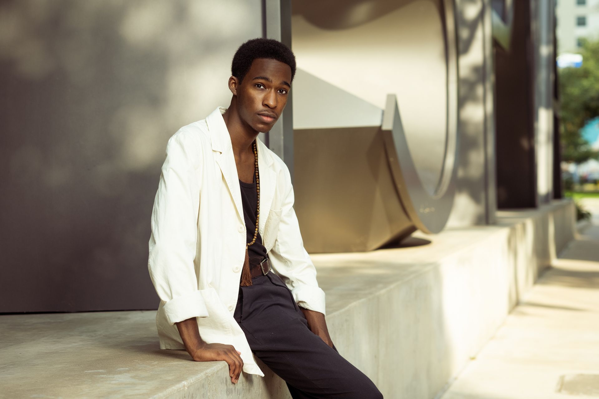Man in white jacket and dark pants sits outdoors, looking at camera.