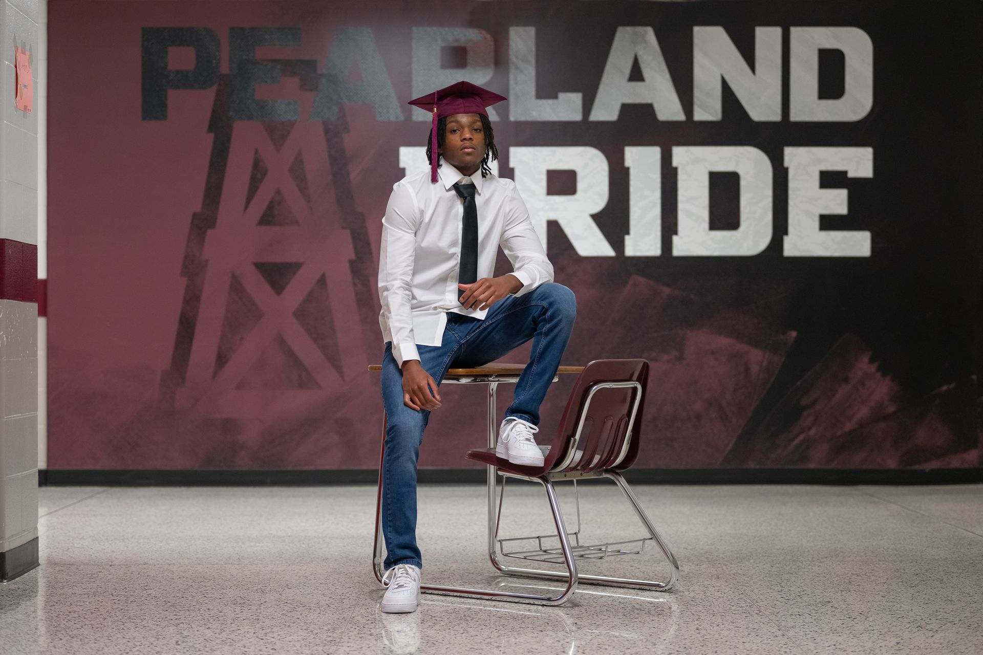 Person in graduation cap poses on a school chair in front of a “Pearland Pride” backdrop.