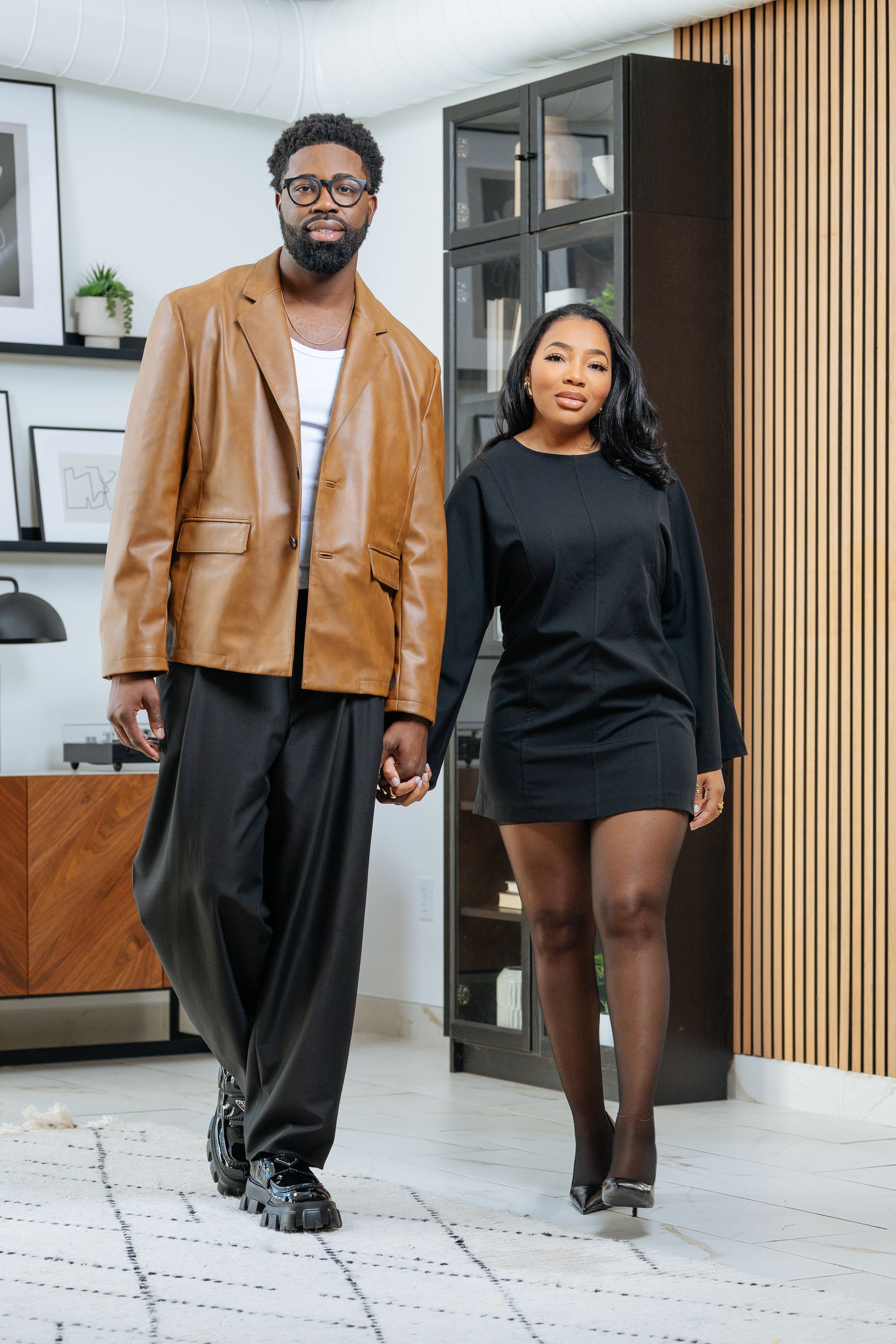 A couple holding hands, walking forward. The man wears a leather jacket, the woman wears a black dress. Modern interior.