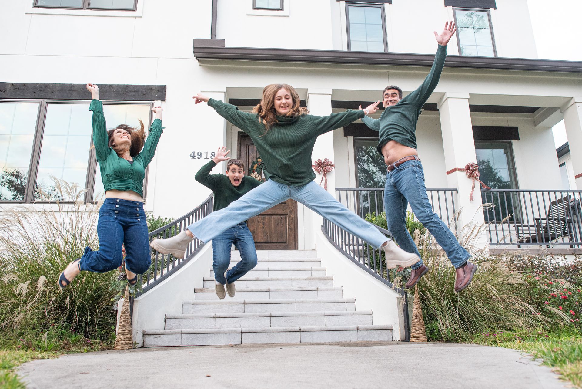 Four people in green shirts and jeans jump in front of a white house with stairs.