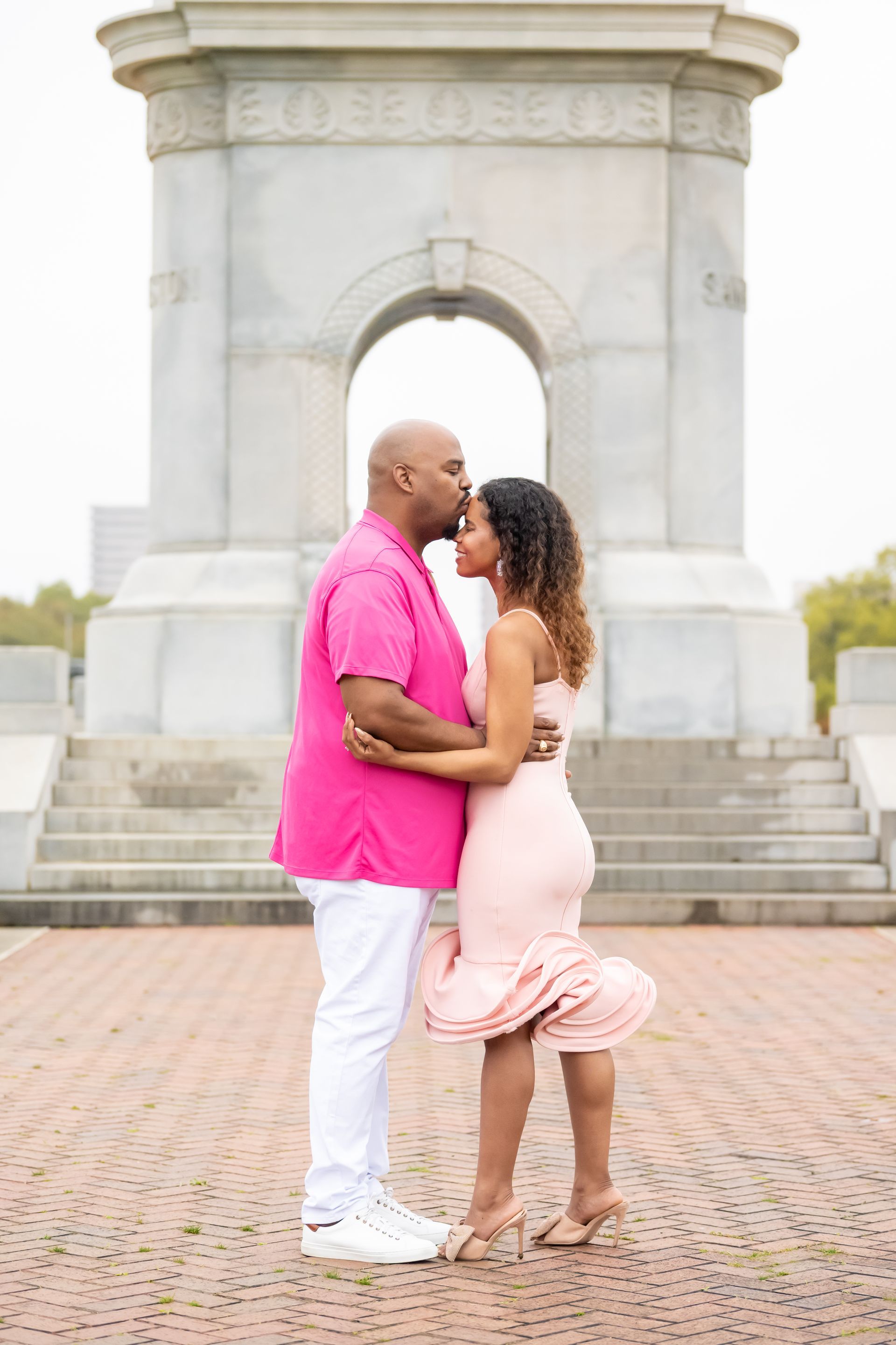 Couple in pink attire embrace, man kissing woman's forehead, in front of monument.