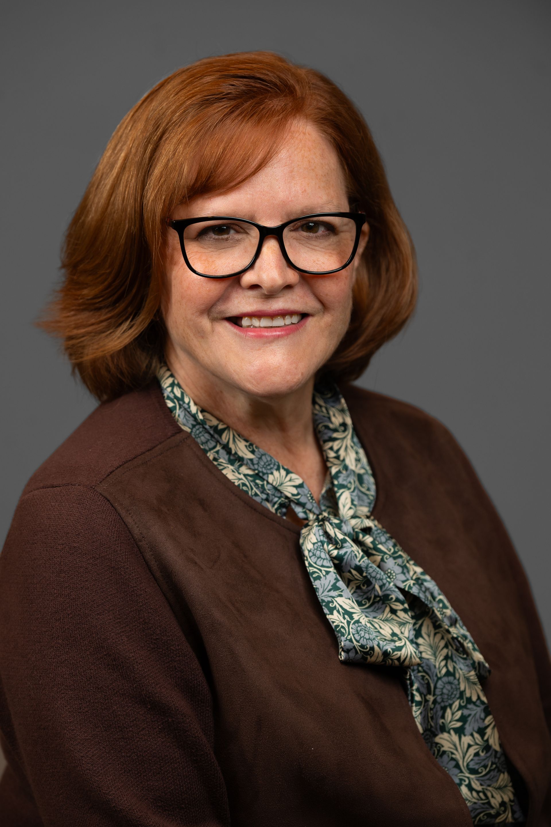 Woman with red hair and glasses smiles, wearing a patterned scarf and brown sweater, against a gray background.