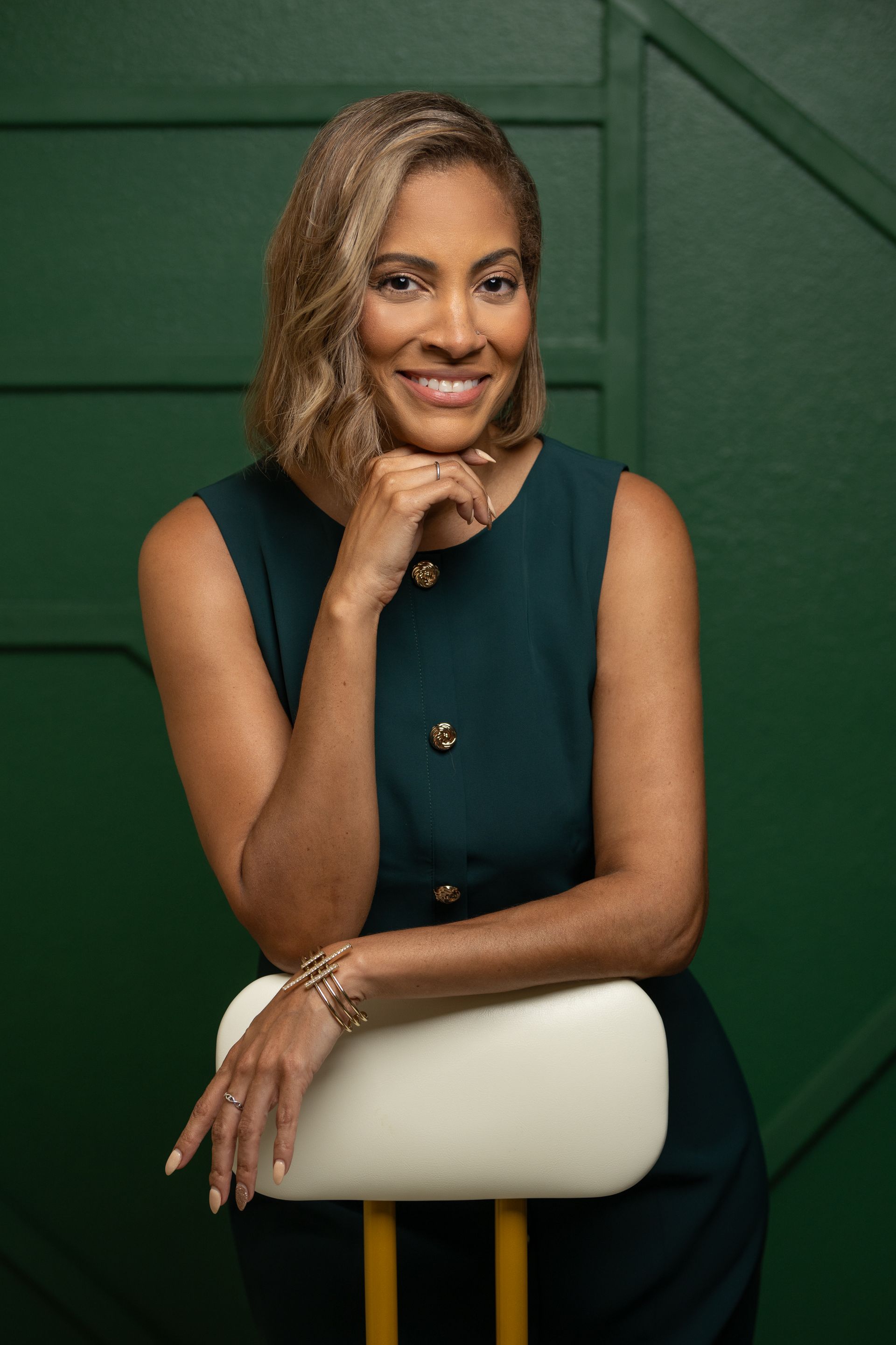 Woman in green dress leans on a chair, smiling, hand on chin, against a green background.