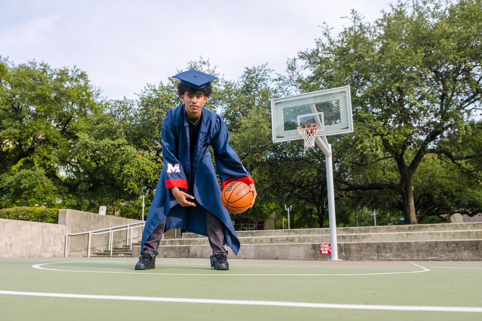 Person in graduation gown dribbling a basketball on a court.