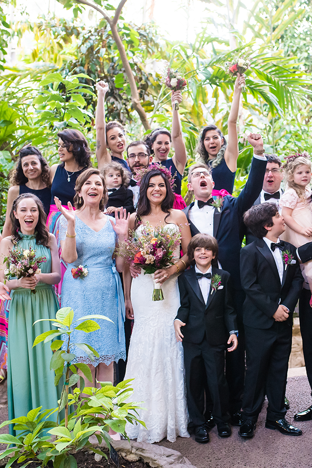 Wedding group in a lush, green setting: Bride with bouquet, guests cheering, some holding flowers.