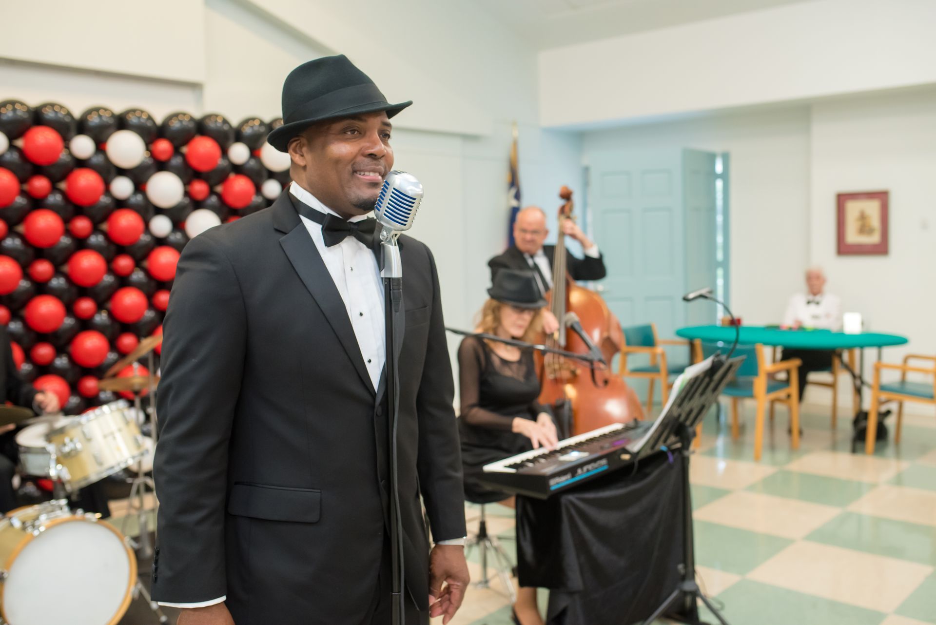 Man in tuxedo singing with band; black, red, and white balloon backdrop.