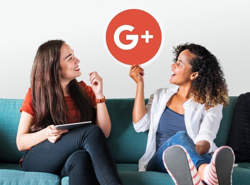 Two women on a teal couch, one holding a Google+ logo, smiling and talking.