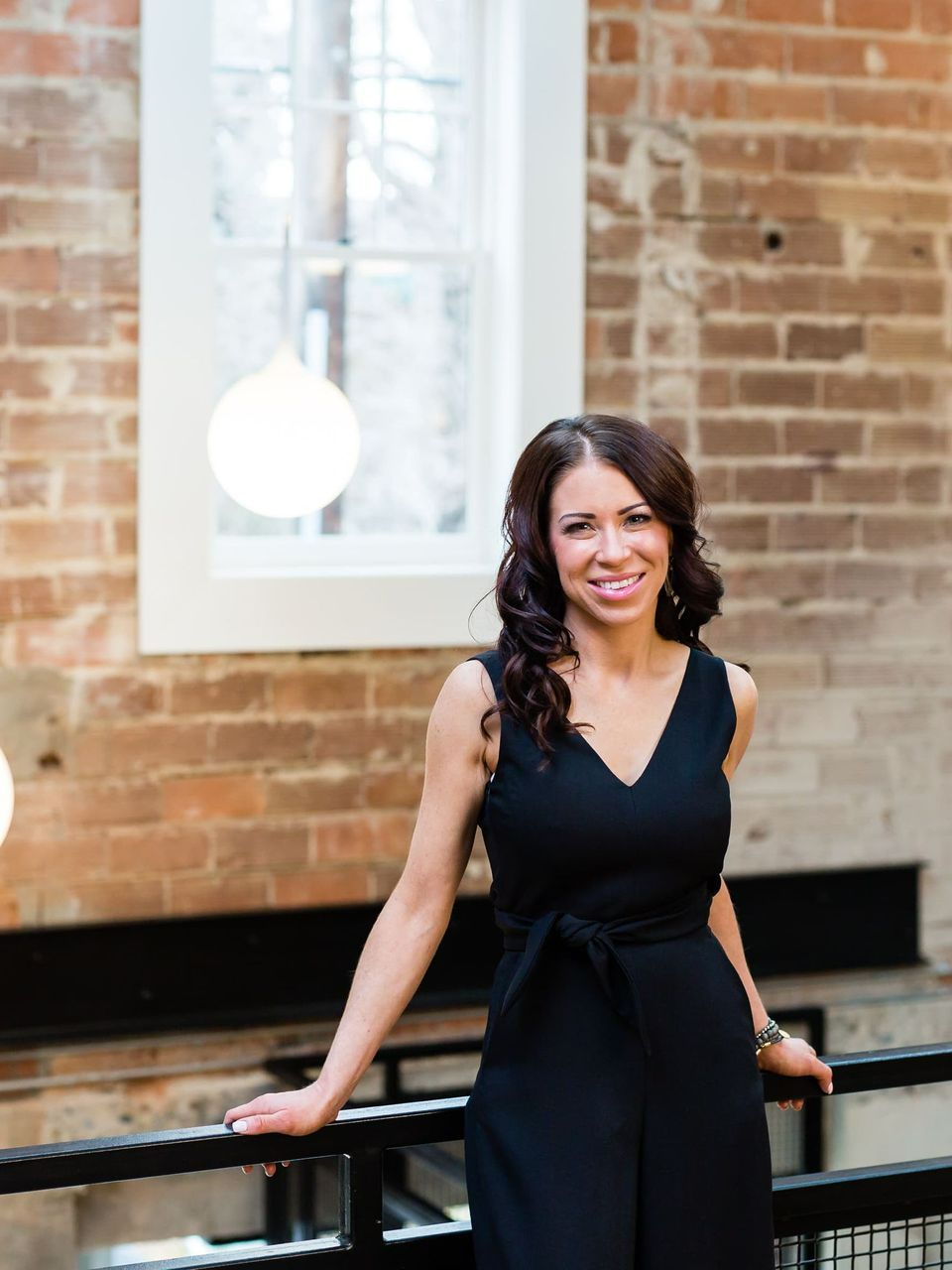 A woman in a black dress is leaning on a railing in front of a brick wall.