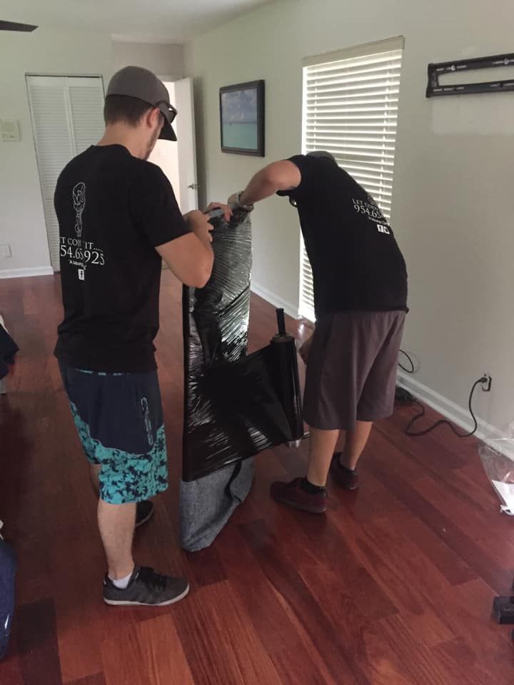 Two people wrapping a tall, black object in a room with hardwood floors