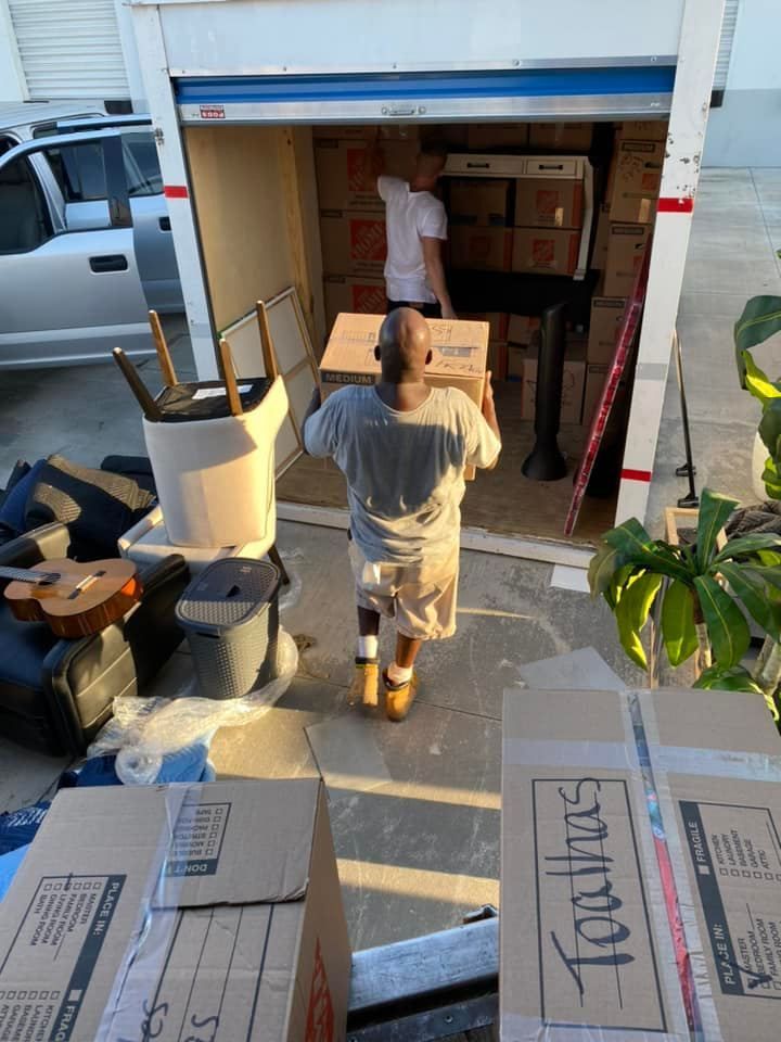 Two people loading boxes into a storage unit. Boxes and furniture are present.