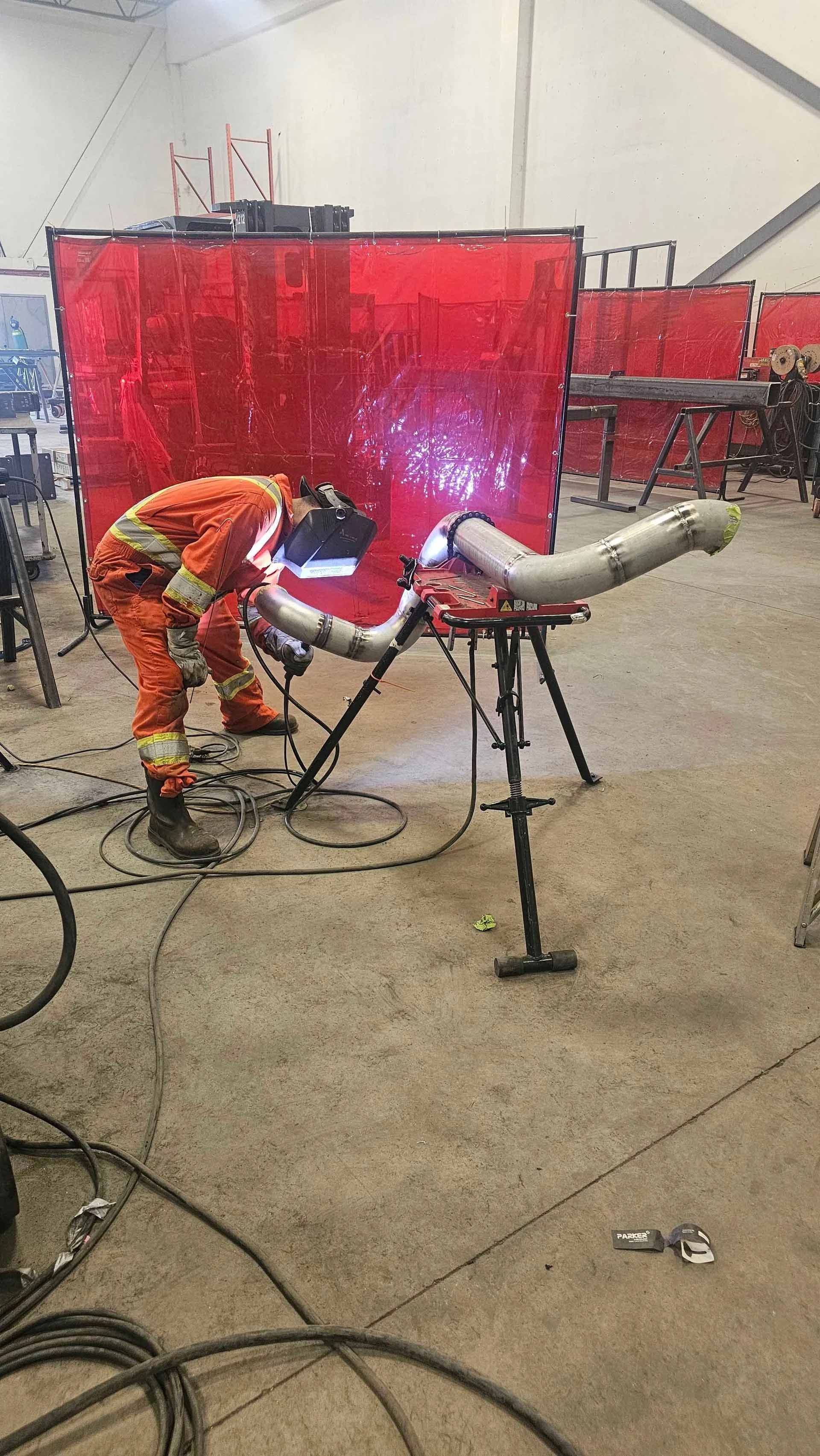 Un soudeur vêtu d'une combinaison de protection orange effectue une soudure à l'arc sur un assemblage de tuyaux métalliques dans un atelier industriel.