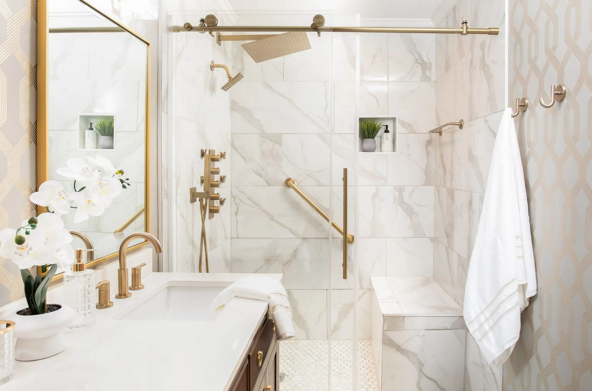 A bright, luxury marble bathroom features a walk-in shower with gold fixtures, a grab bar, a tiled bench, and a vanity.