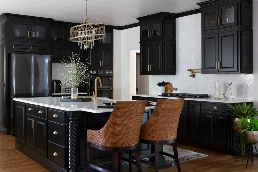 A black-cabinet kitchen with a white island, gold hardware, two woven bar stools, and a gold chandelier.