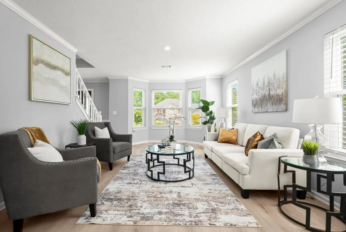 A bright, airy living room with a white sofa, two gray armchairs, a geometric coffee table, and large bay windows.