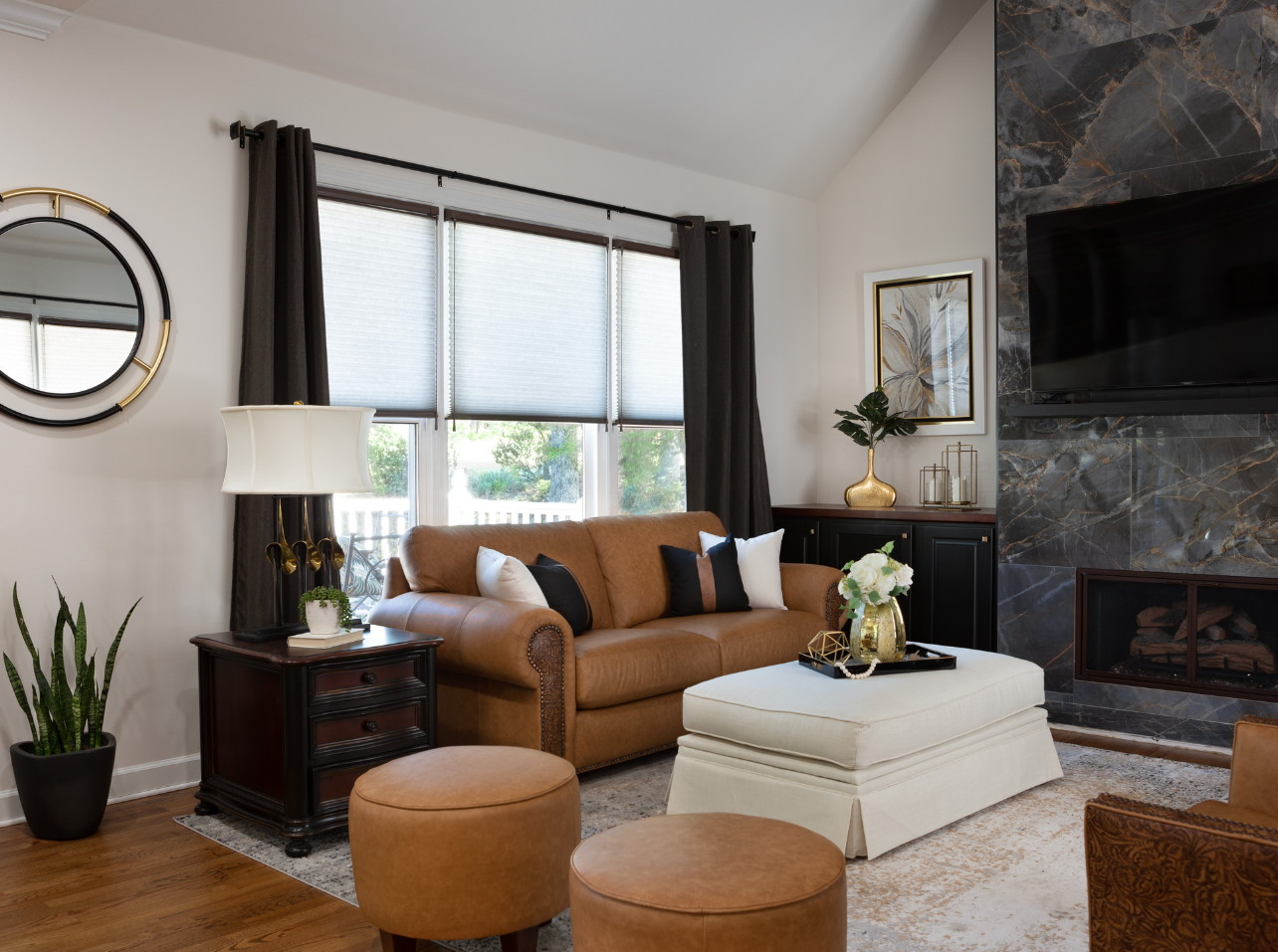 A cozy living room with a tan sofa, cream ottoman, two round leather stools, and a fireplace with a mounted television.
